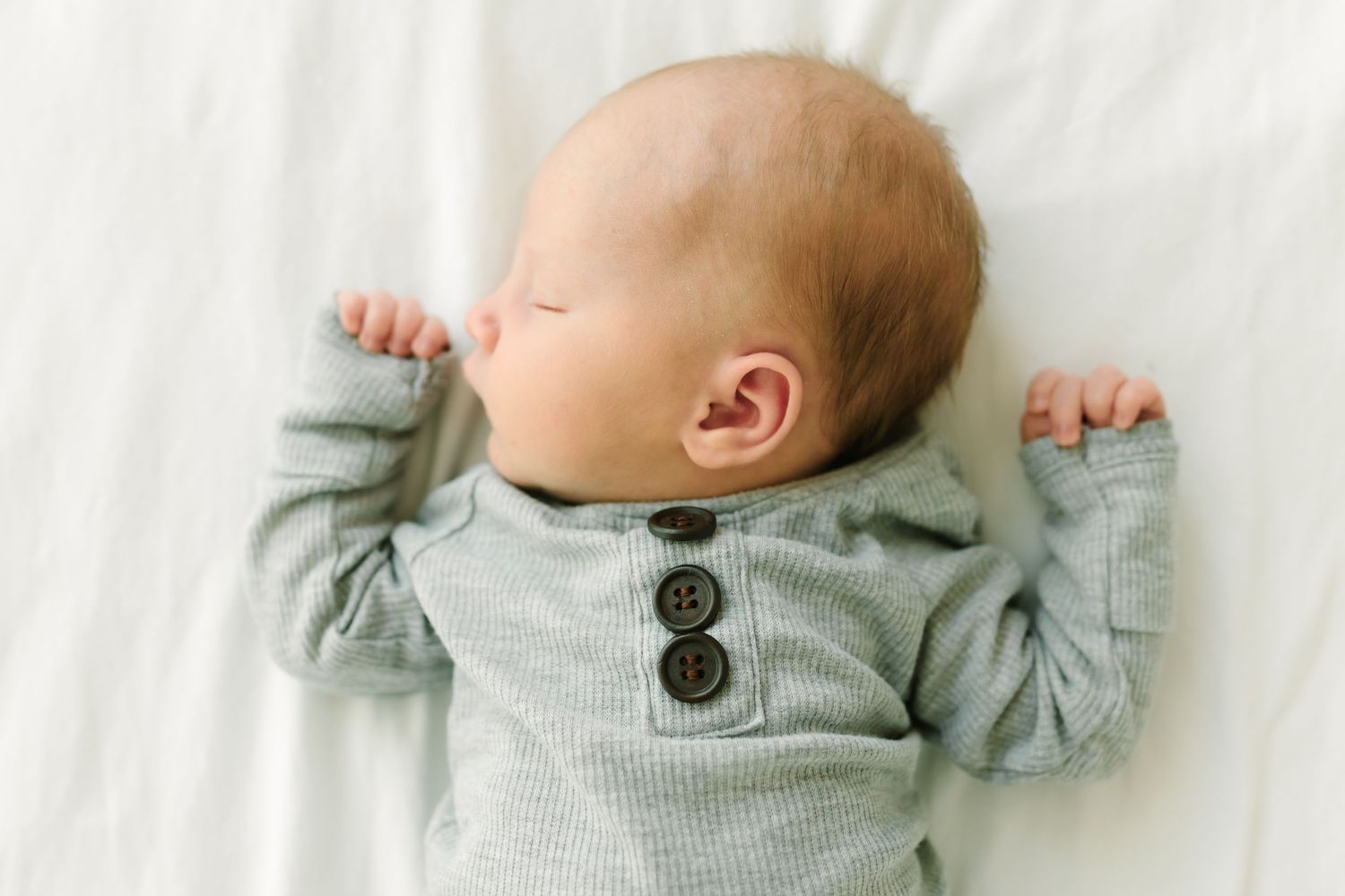 Peaceful newborn baby sleeping on white blanket wearing a soft gray ribbed button onesie.