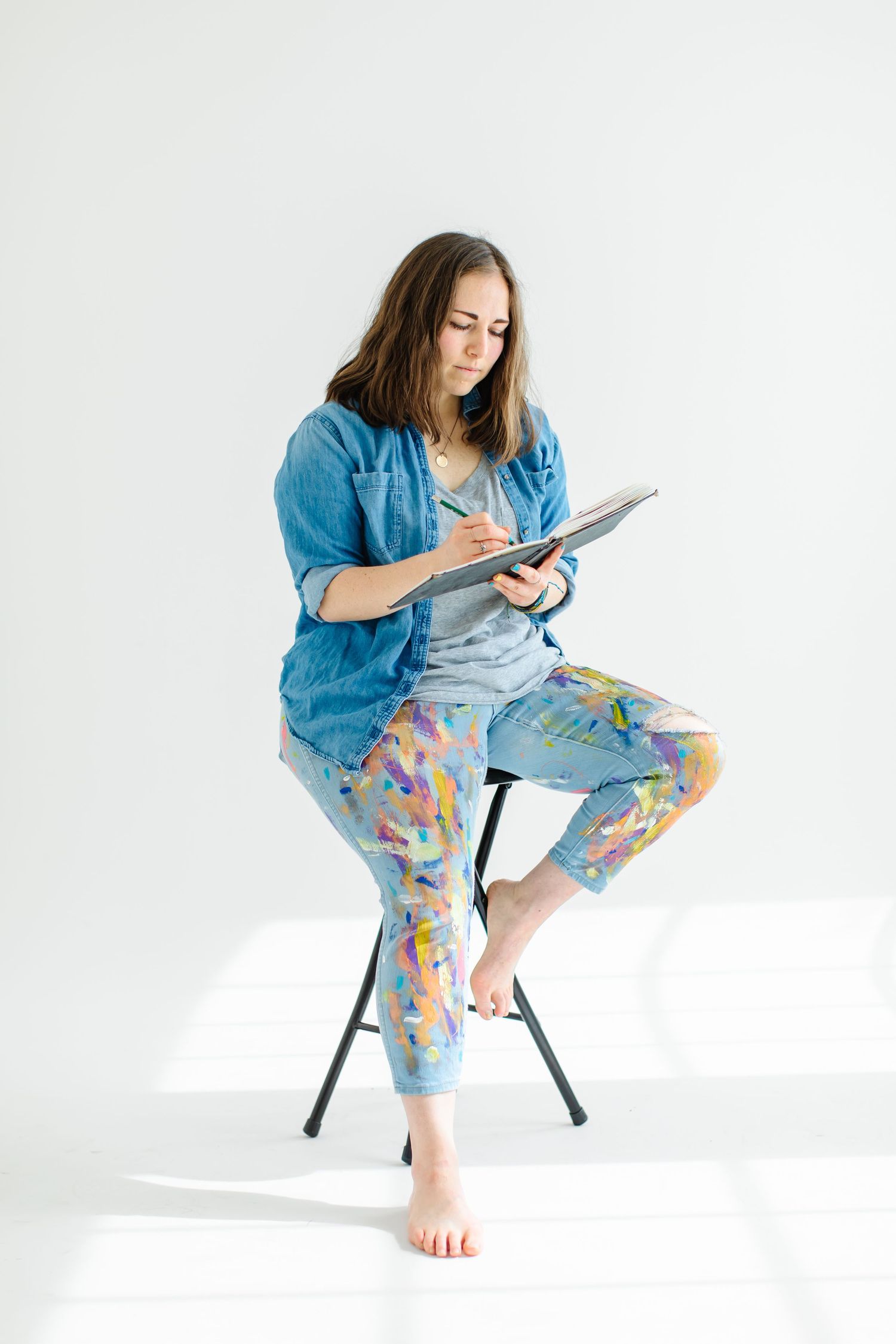 Artist sketches while sitting on stool wearing colorful pants and denim shirt at White Space Studios in Provo, Utah in Utah County..