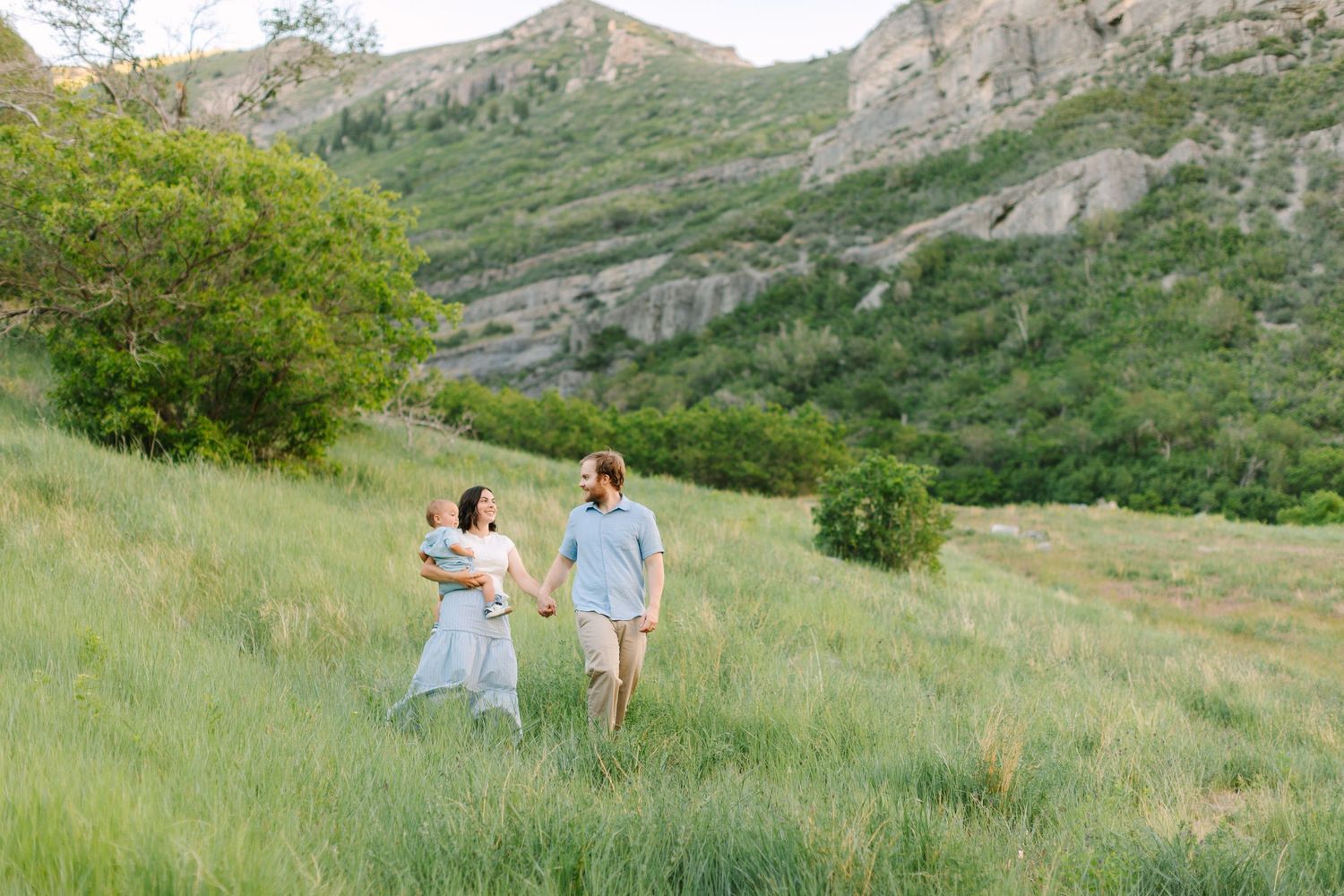 A couple walks hand-in-hand through a lush green meadow with rocky mountain slopes in the background in Provo, Utah.