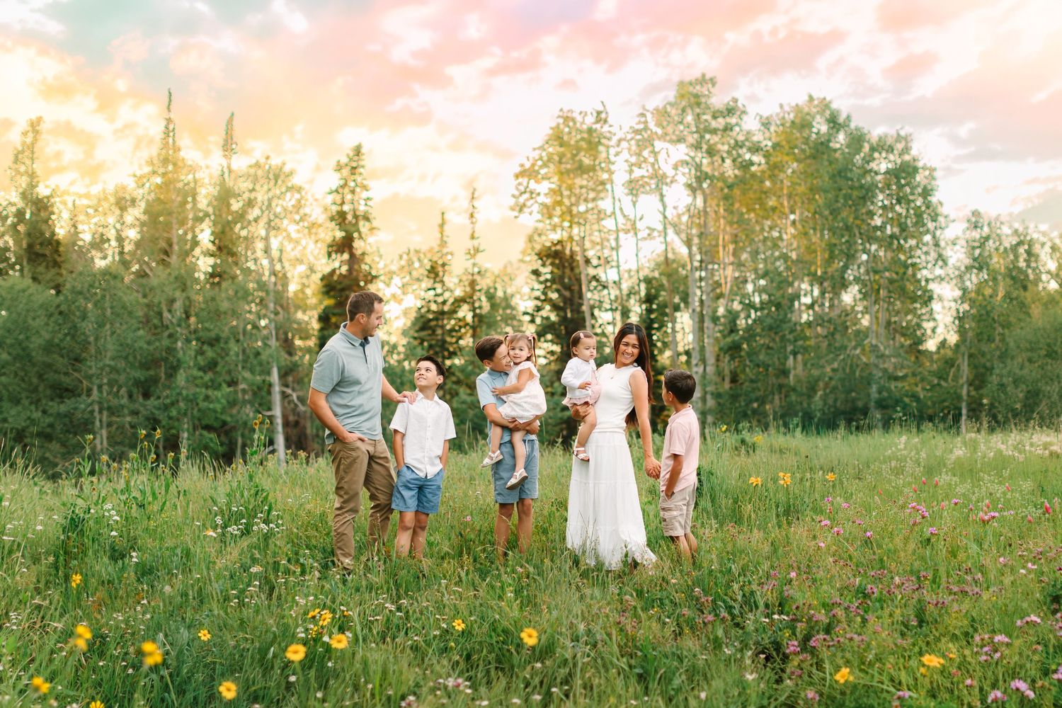 Family of five standing in a wildflower meadow at golden hour with lush green trees and a colorful sunset sky.