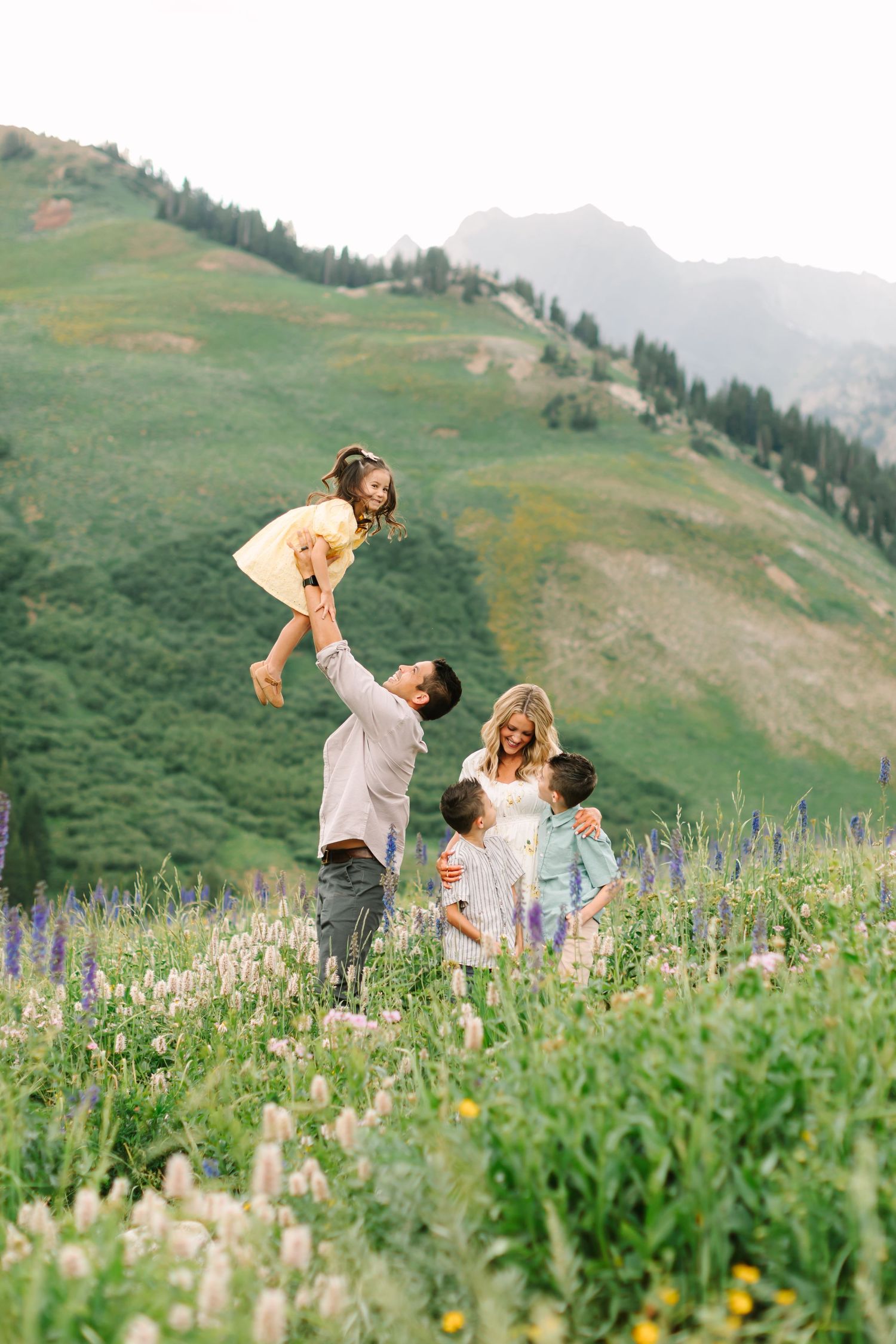 Family playing in a wildflower meadow with mountains, dad lifting daughter while mom hugs two sons.