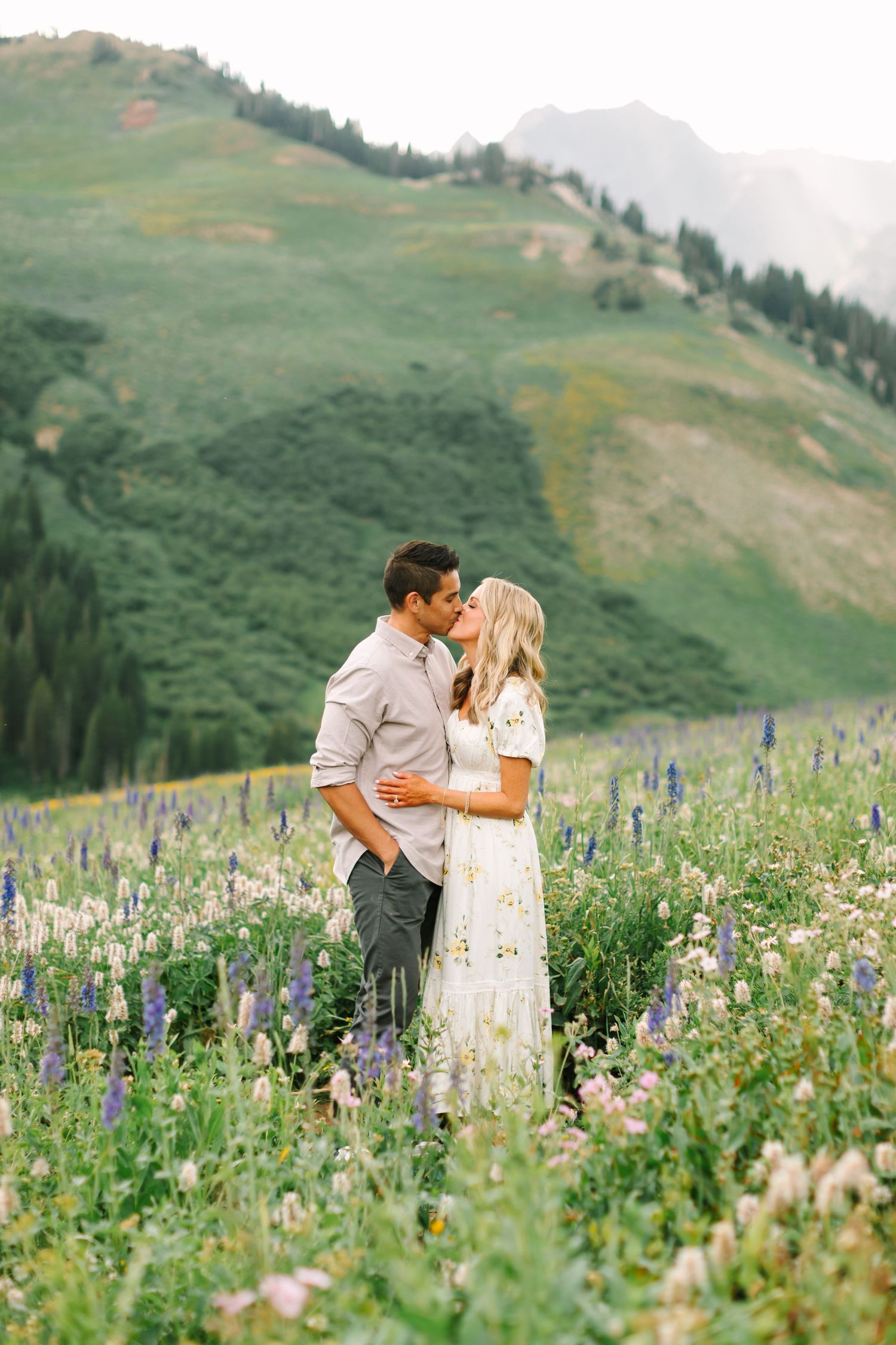 Couple kissing in a wildflower meadow with purple lupines, green hills, and misty mountains in the background.