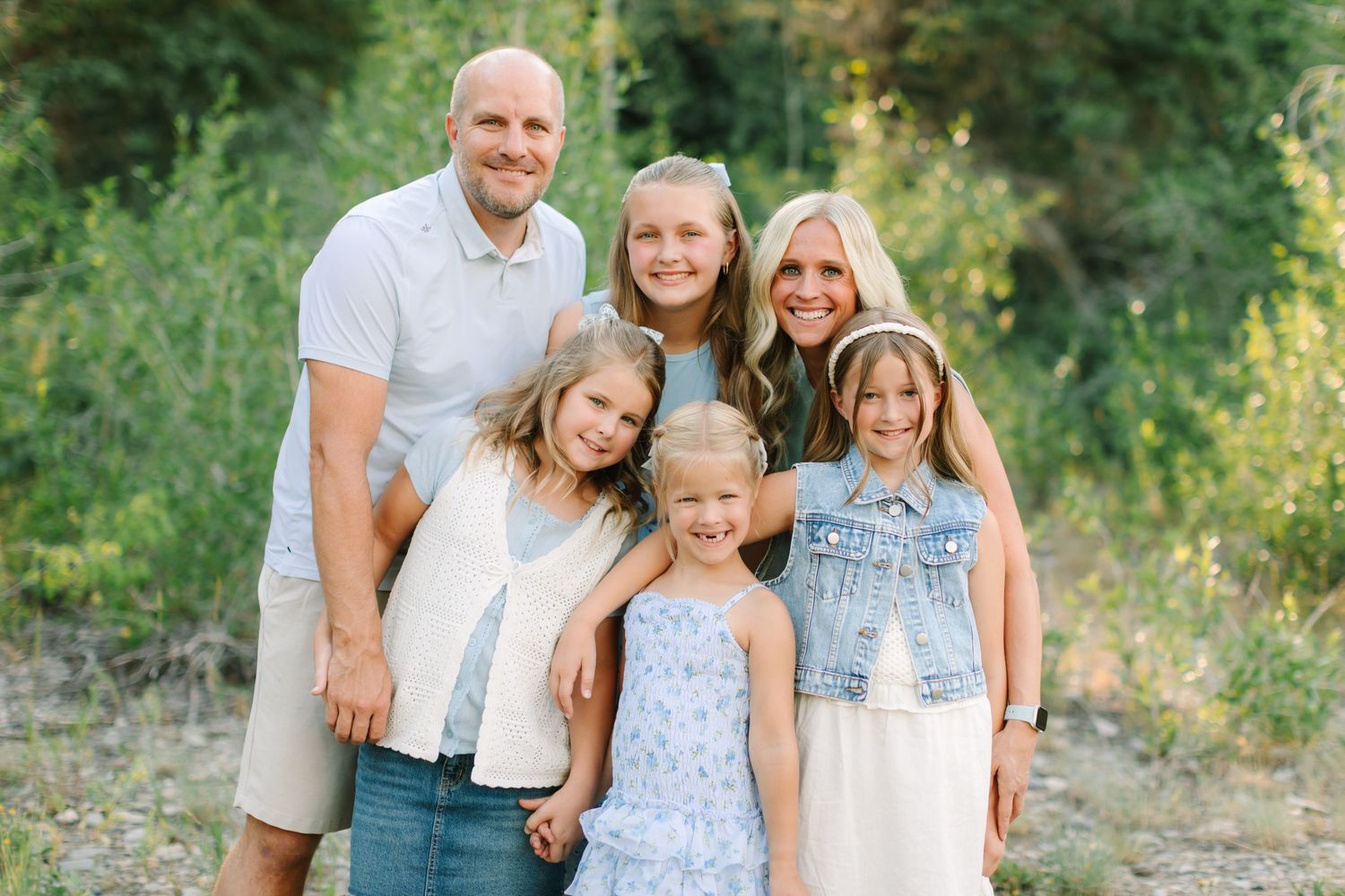 Large family group smiling outdoors in coordinated sage green and white outfits with lush green trees behind in Provo, Utah.