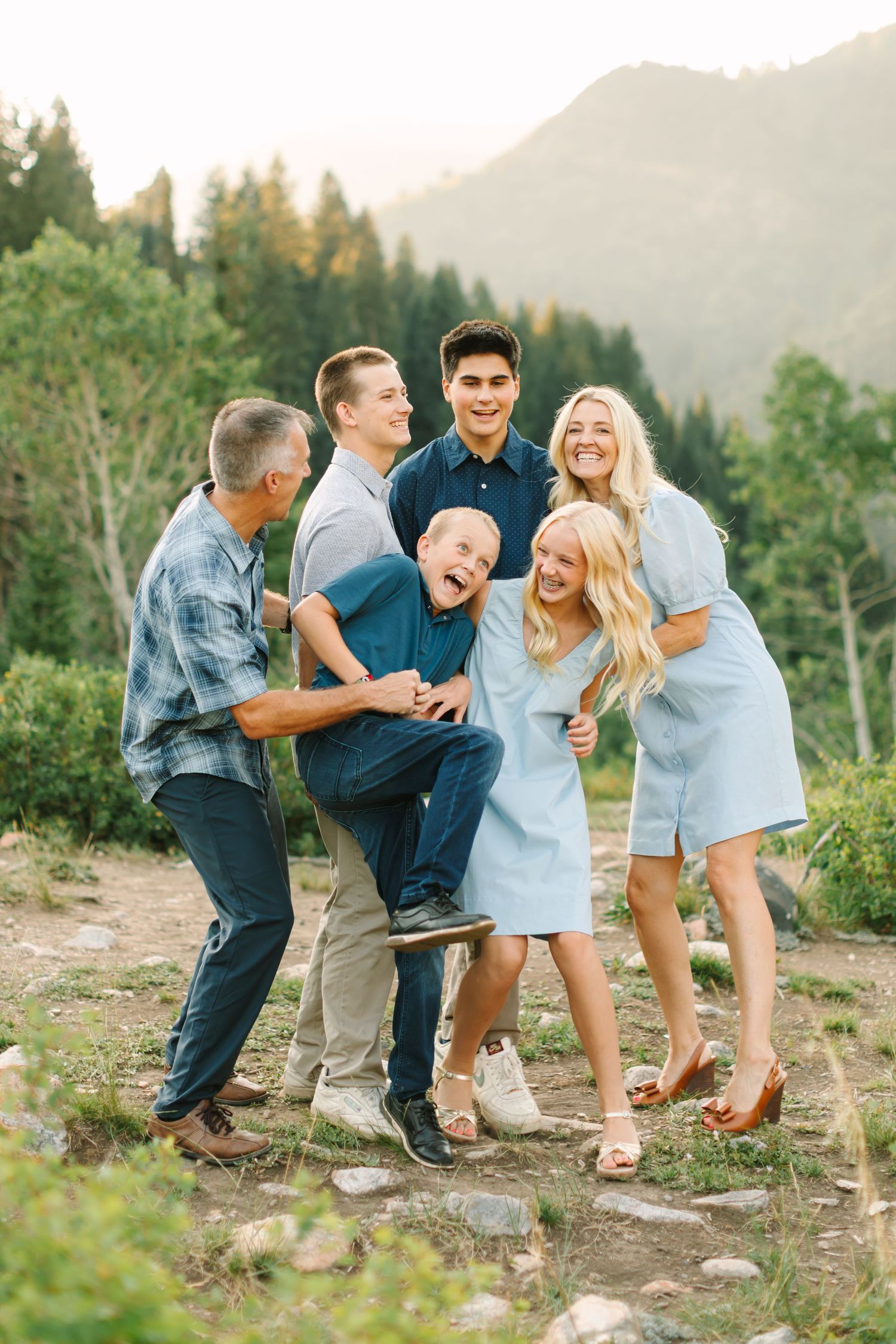 A joyful family of seven laughing together outdoors in a mountain setting, dressed in coordinating blue outfits.