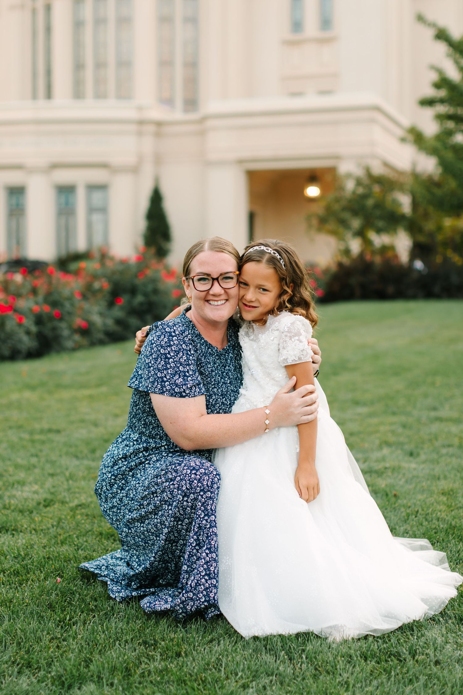 Two people share a tender embrace while sitting on lush green grass near a grand white building.