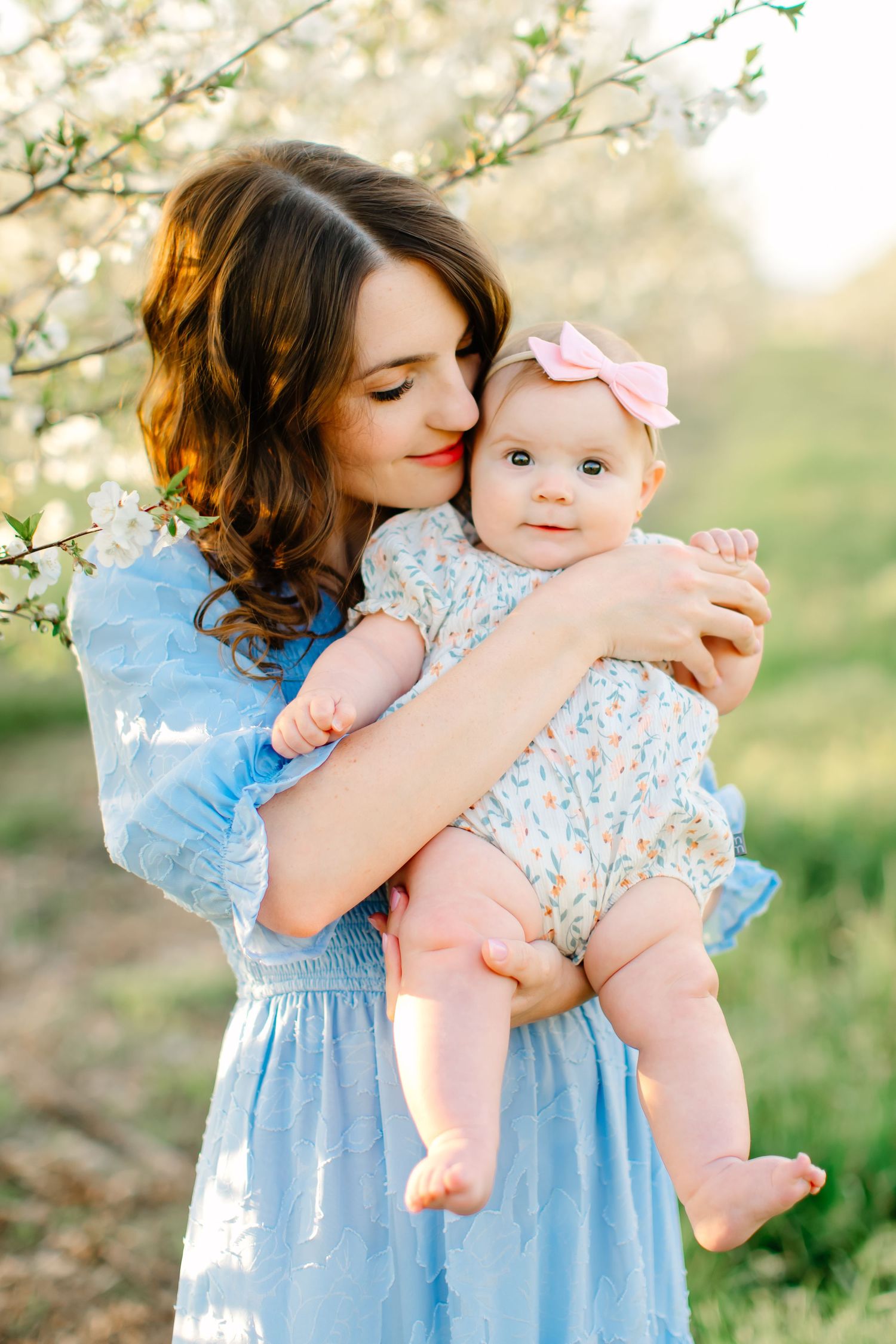 A tender moment captured in spring as a mother in a blue dress embraces a baby wearing a floral outfit among blooming trees in Santaquin, Ut