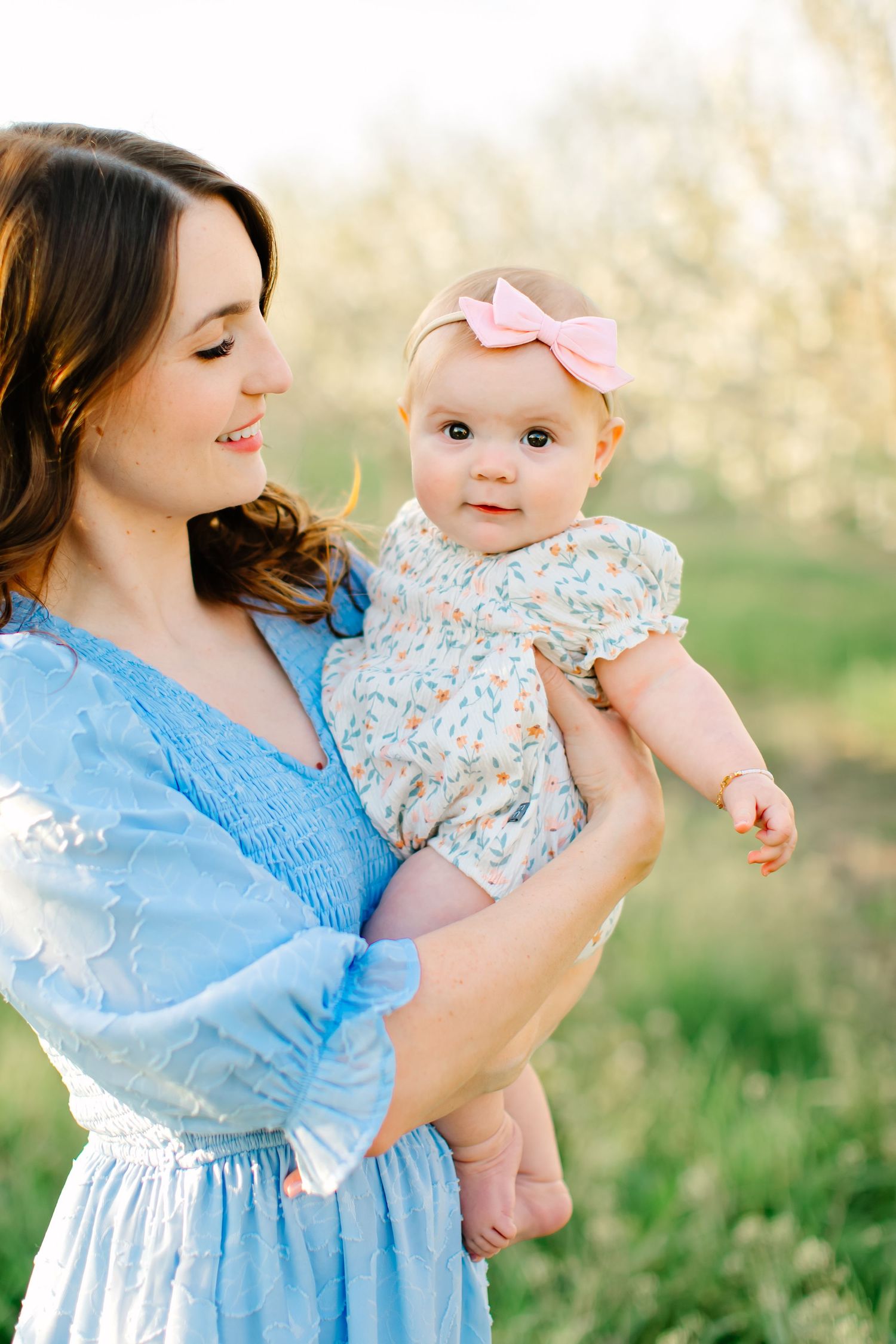 A mother in a blue dress holds a baby wearing a floral outfit in a sunny outdoor setting in Santaquin, Utah.
