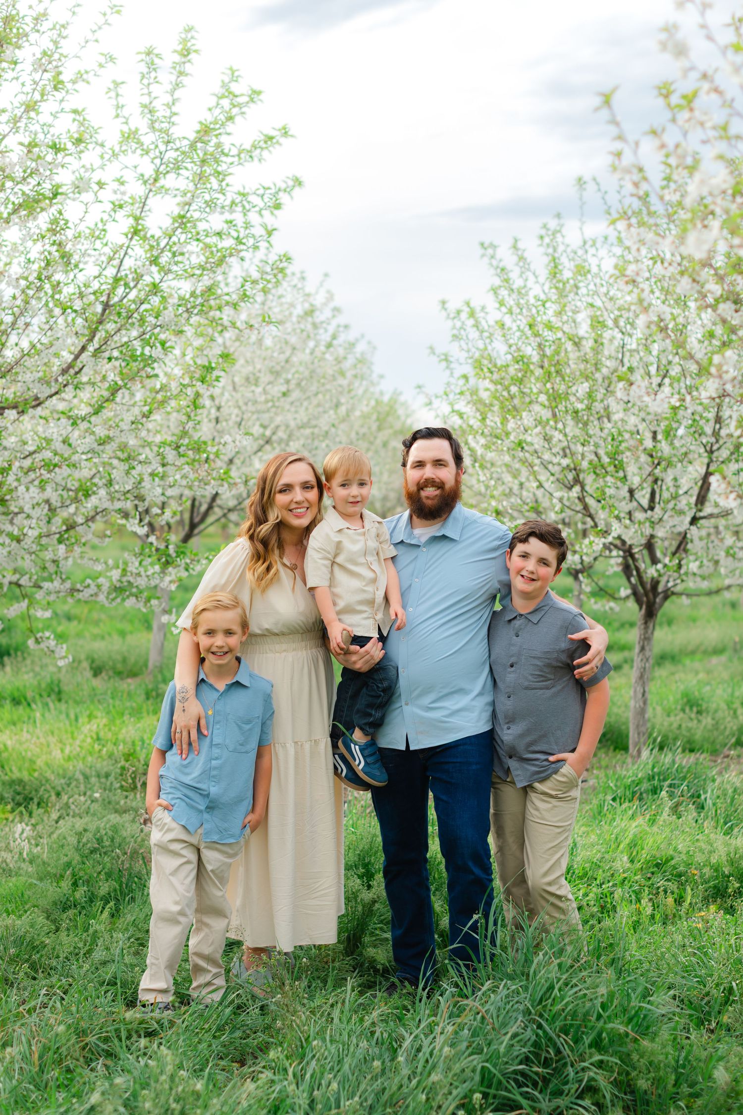 A family poses together in a blooming spring orchard with white flowering trees and lush green grass.