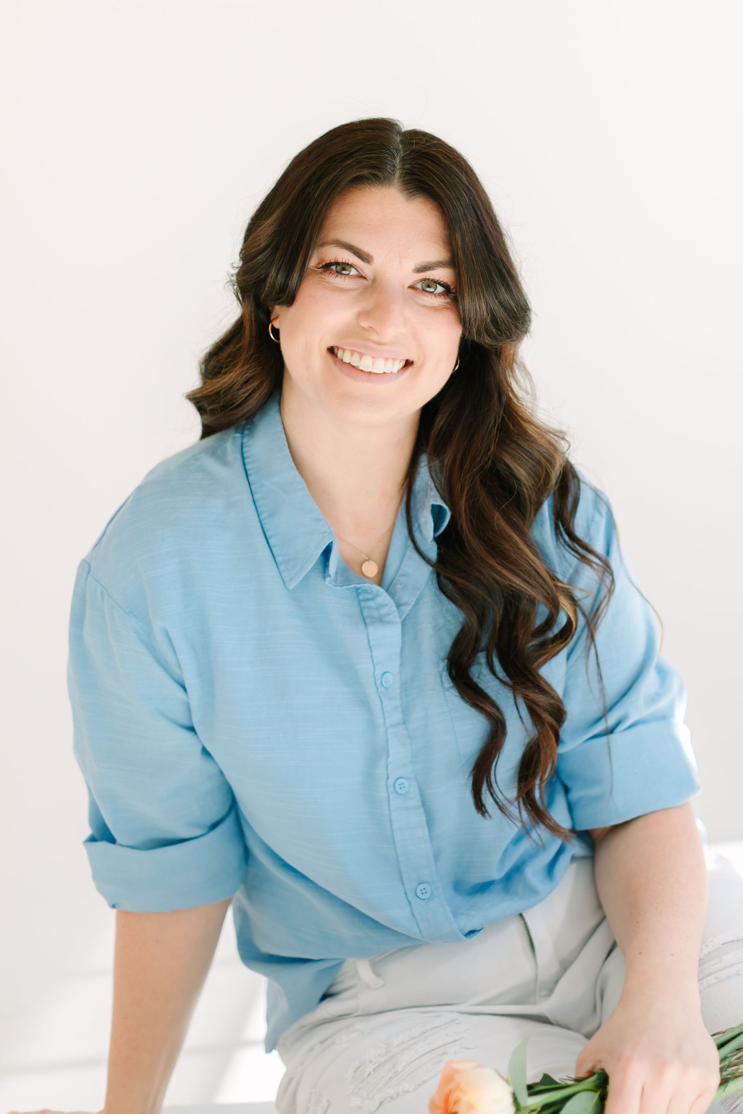 Professional portrait of someone in a light blue button-down shirt with long dark wavy hair against white background.