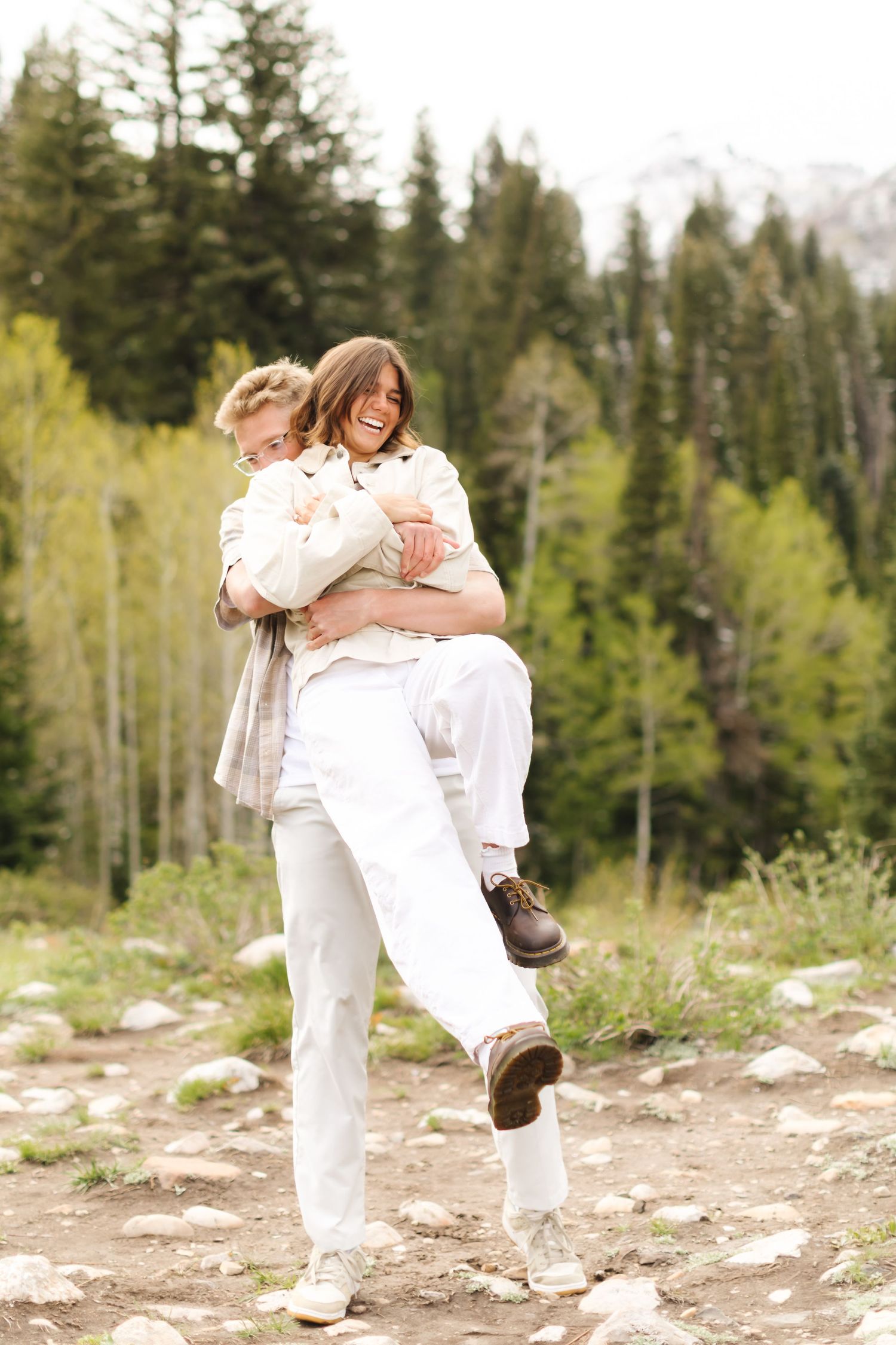 Couple in white outfits laughing outdoors, man carrying woman piggyback on rocky trail with green forest background.