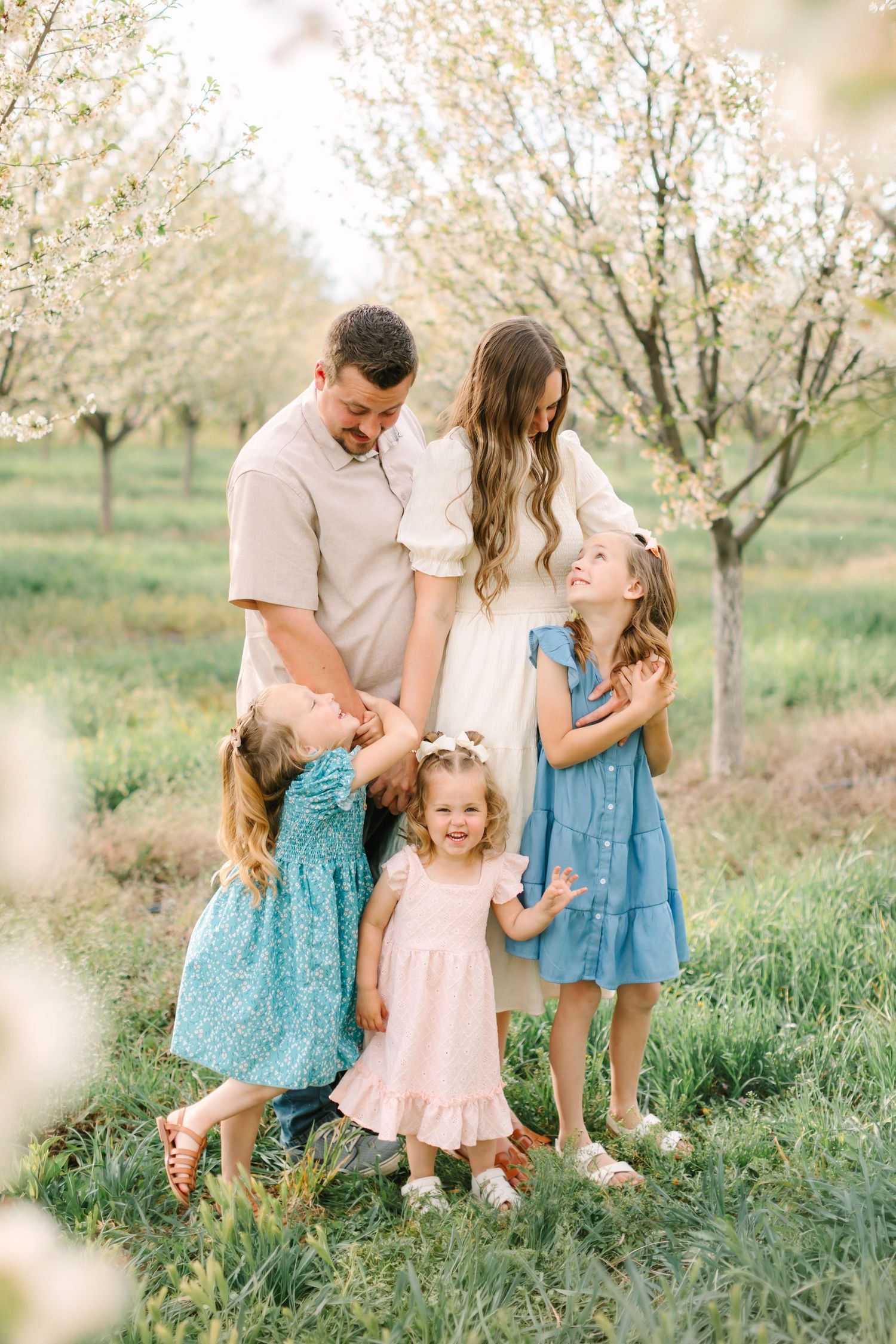 Family of five posing in blooming orchard, parents embracing with three young daughters in pastel dresses on green grass.