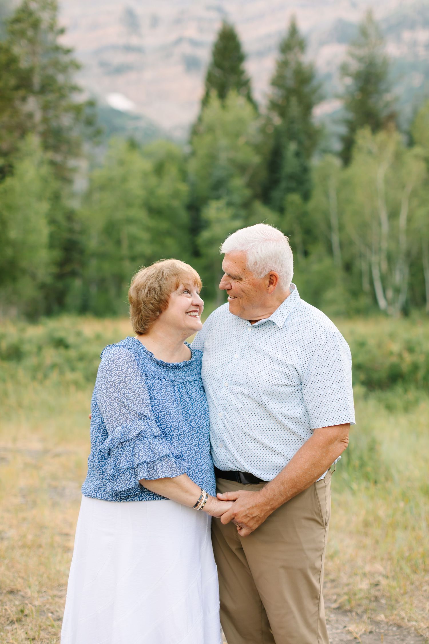 Elderly couple smiling at each other outdoors, surrounded by green trees and mountains in the background.