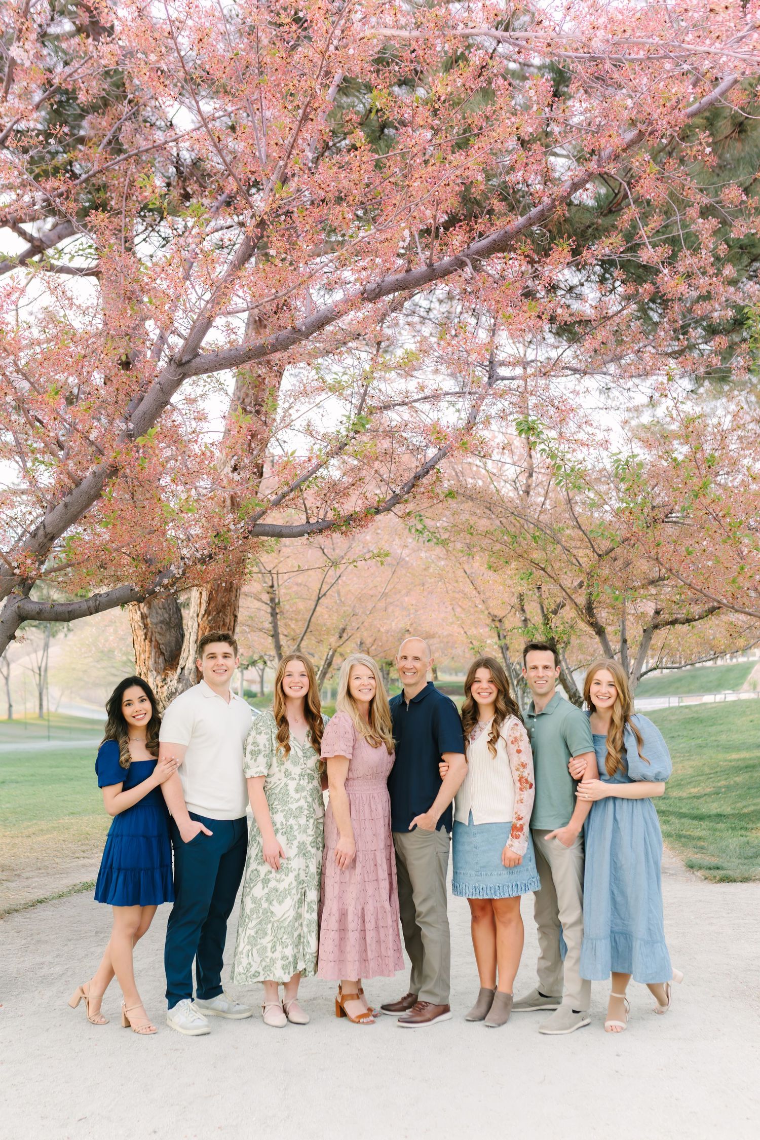 A family of eight poses under blooming pink cherry blossom trees, dressed in coordinated spring outfits.