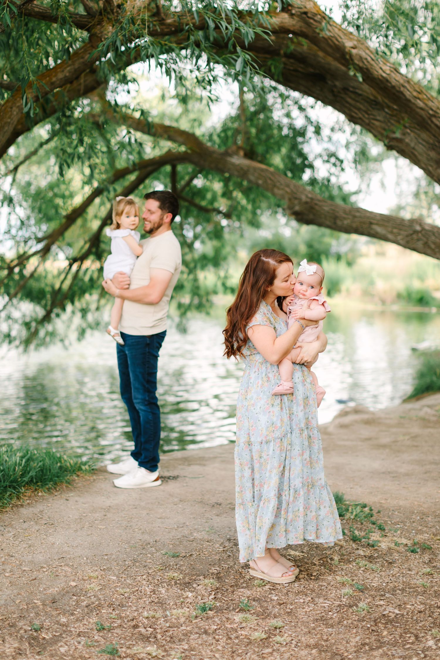A family poses by a serene lake under a willow tree, parents each holding a young child outdoors.