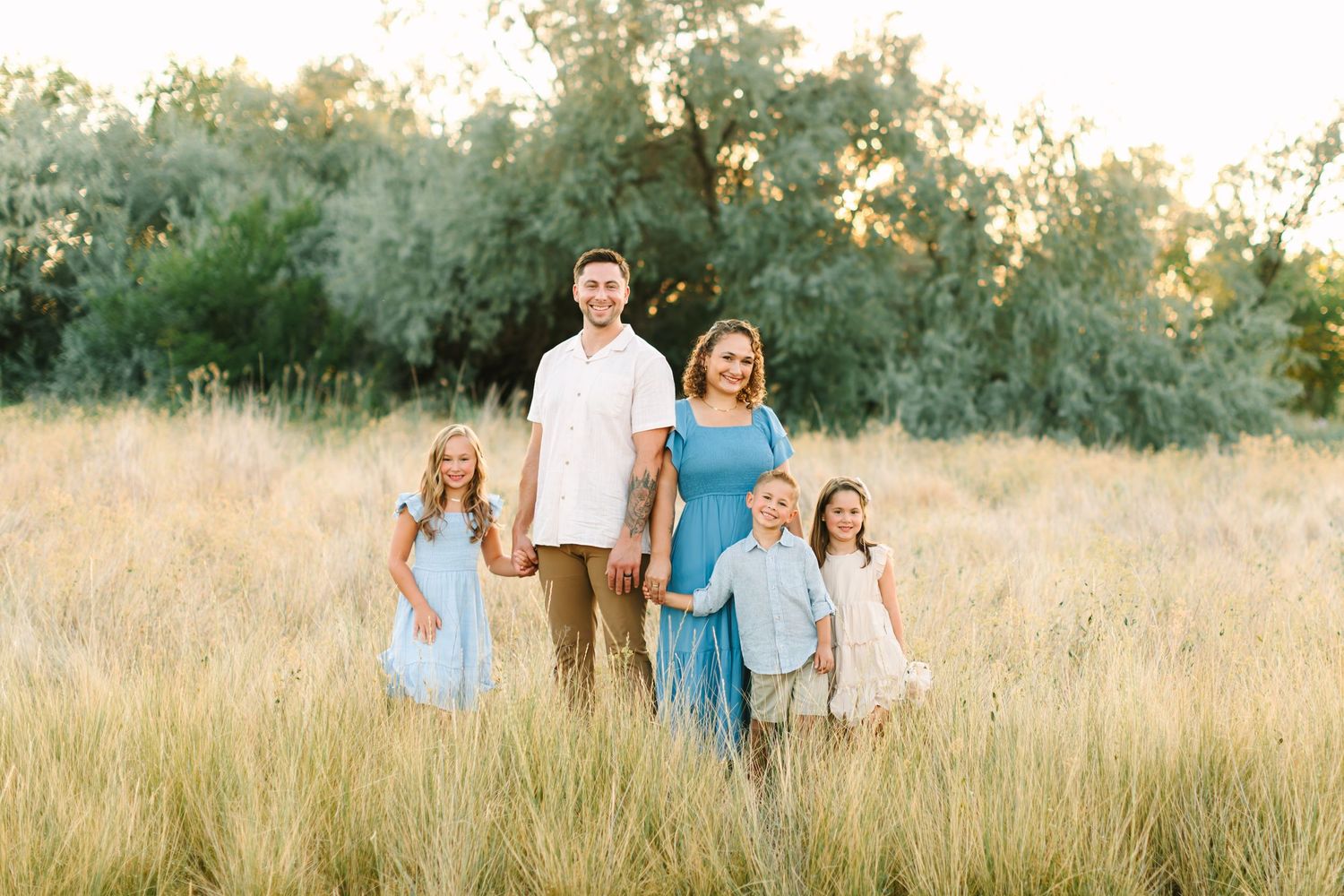 Family of five posing in golden field with tall grass and trees, wearing coordinated blue and white outfits in Bountiful, Utah.