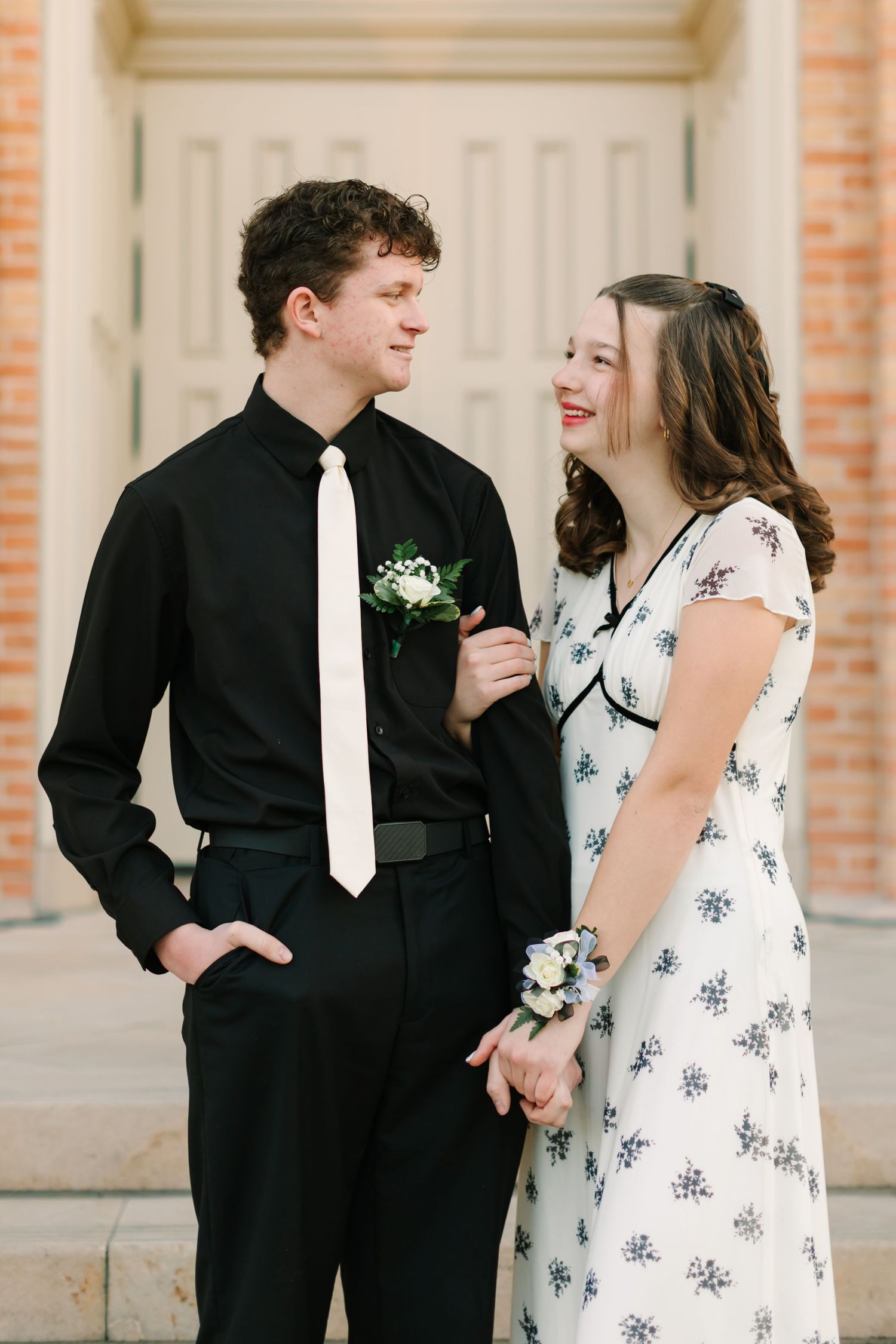 A couple in formal attire share a joyful moment at prom, with her in a floral dress and him in black with a white tie.