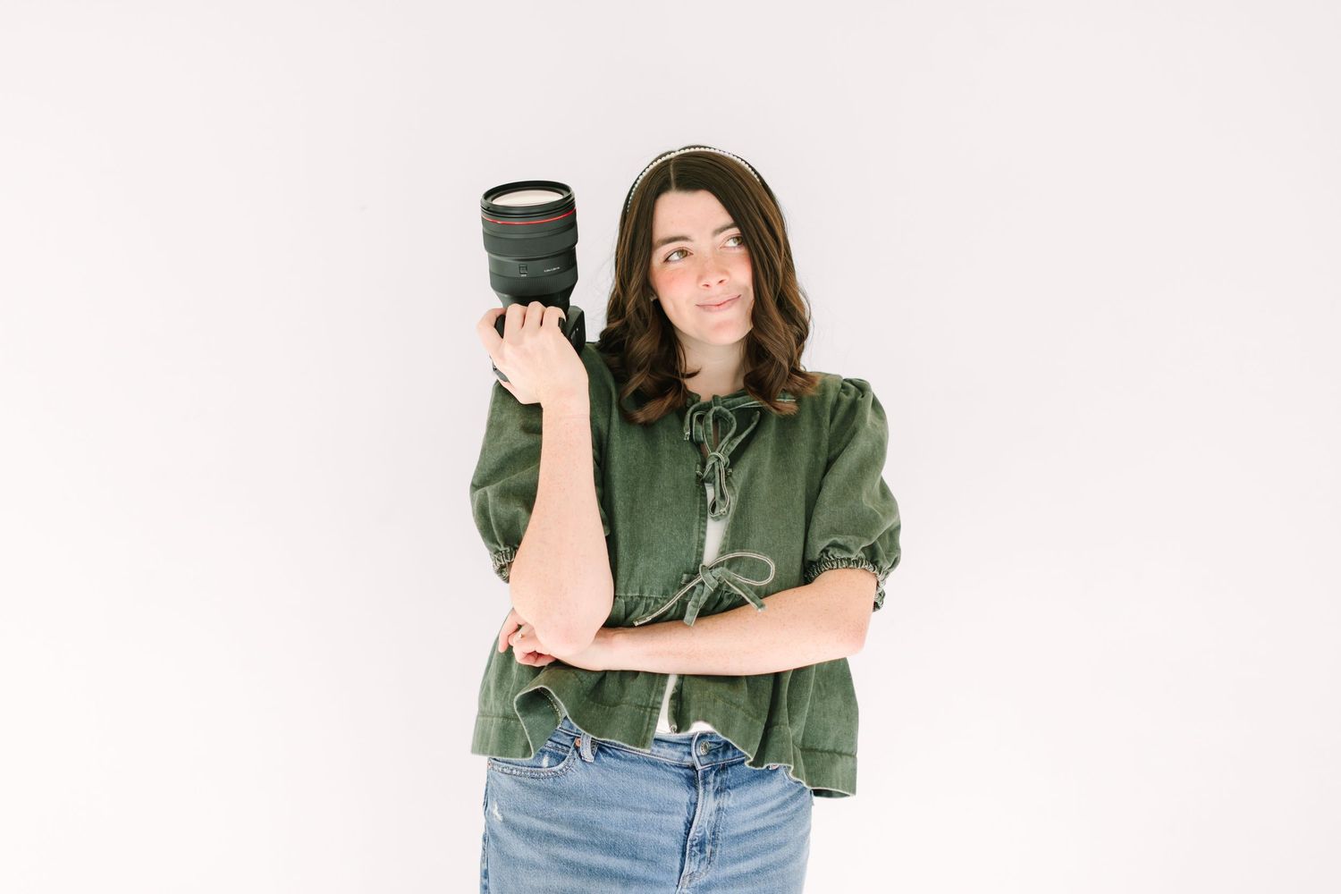 Person in a green button-up shirt and jeans holding a camera while posing against a white background.