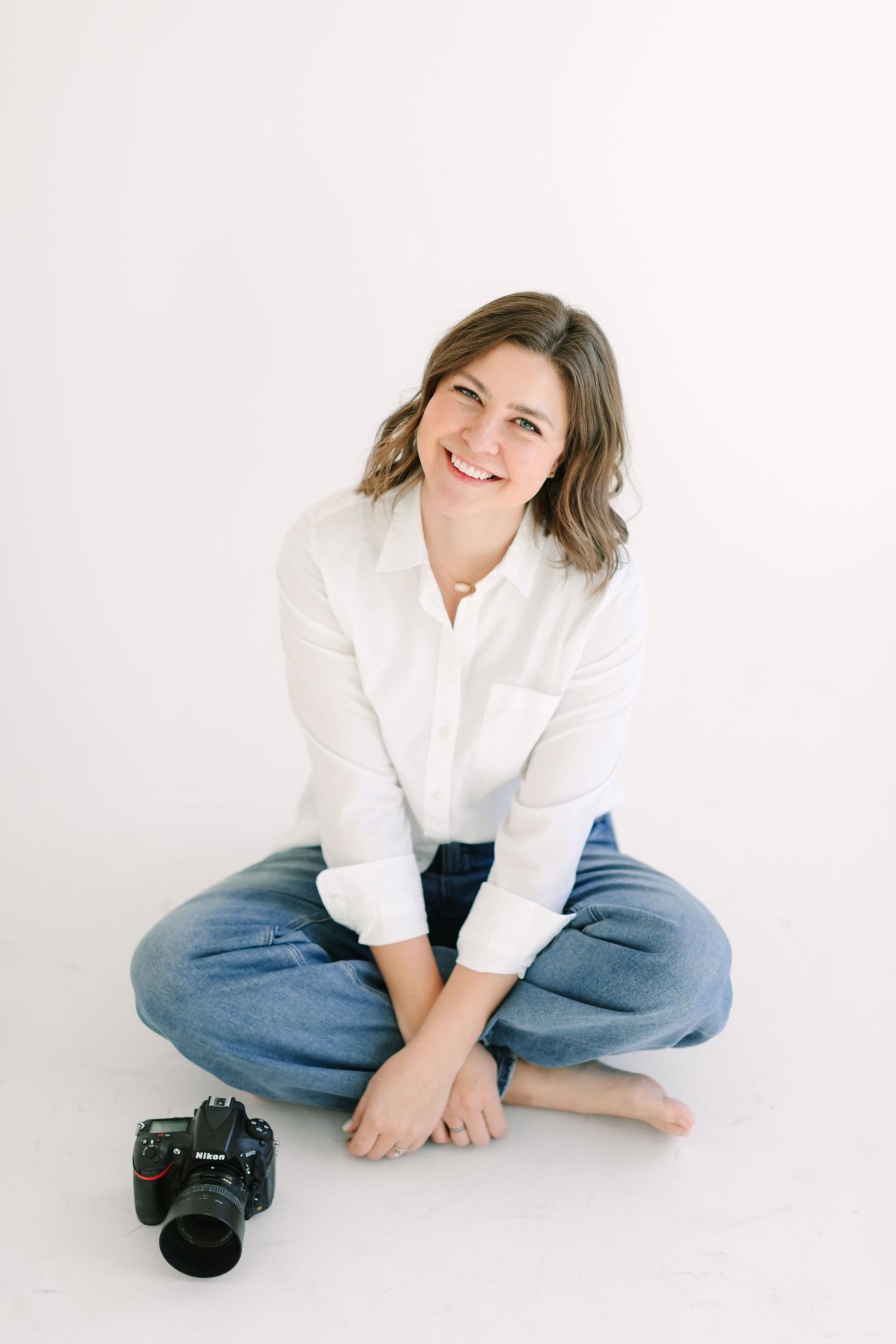 Professional photographer in white shirt and jeans sits cross-legged on floor with camera beside them on white background.