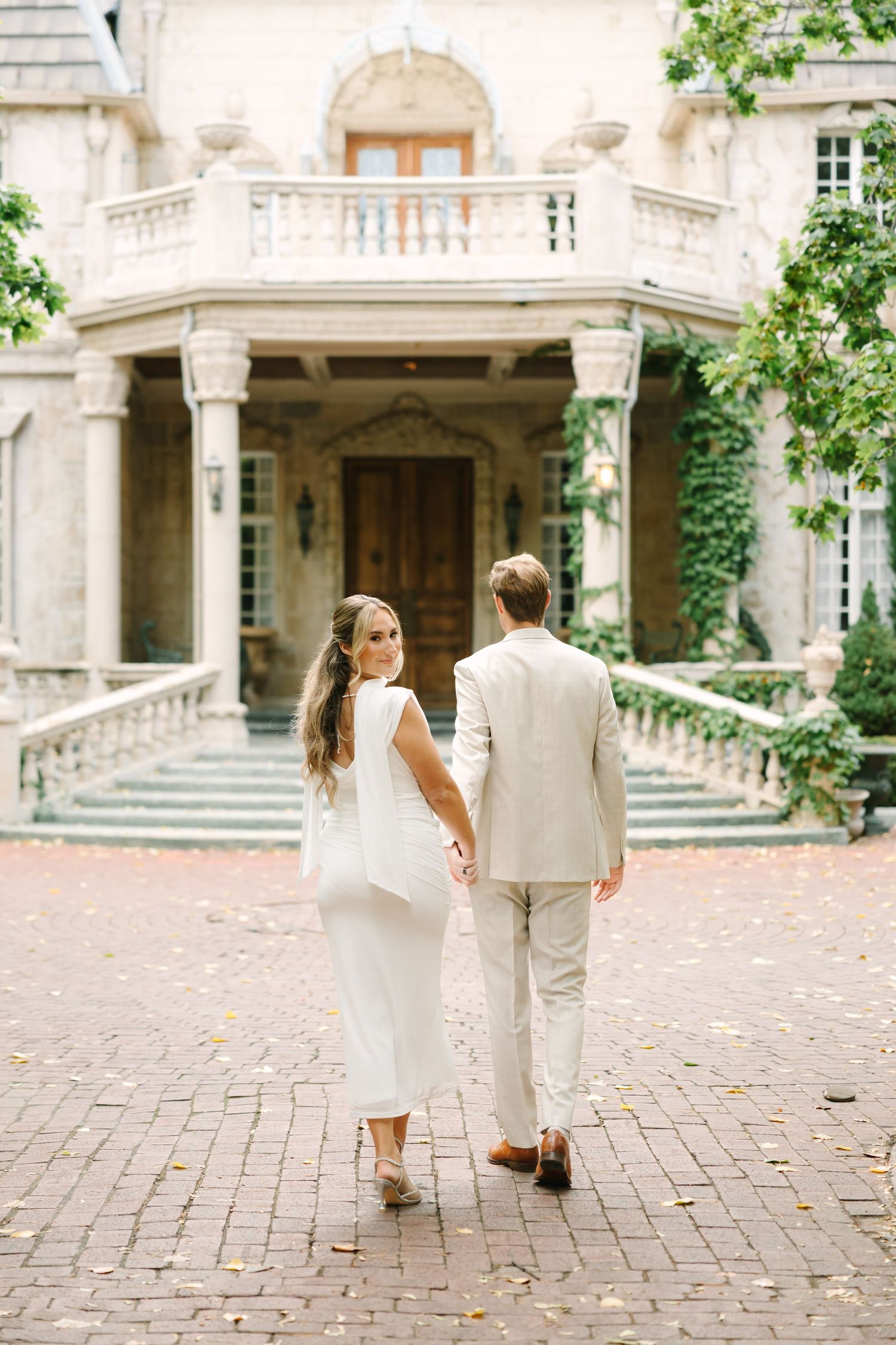 A couple walks hand in hand on a brick courtyard surrounded by elegant stone architecture.