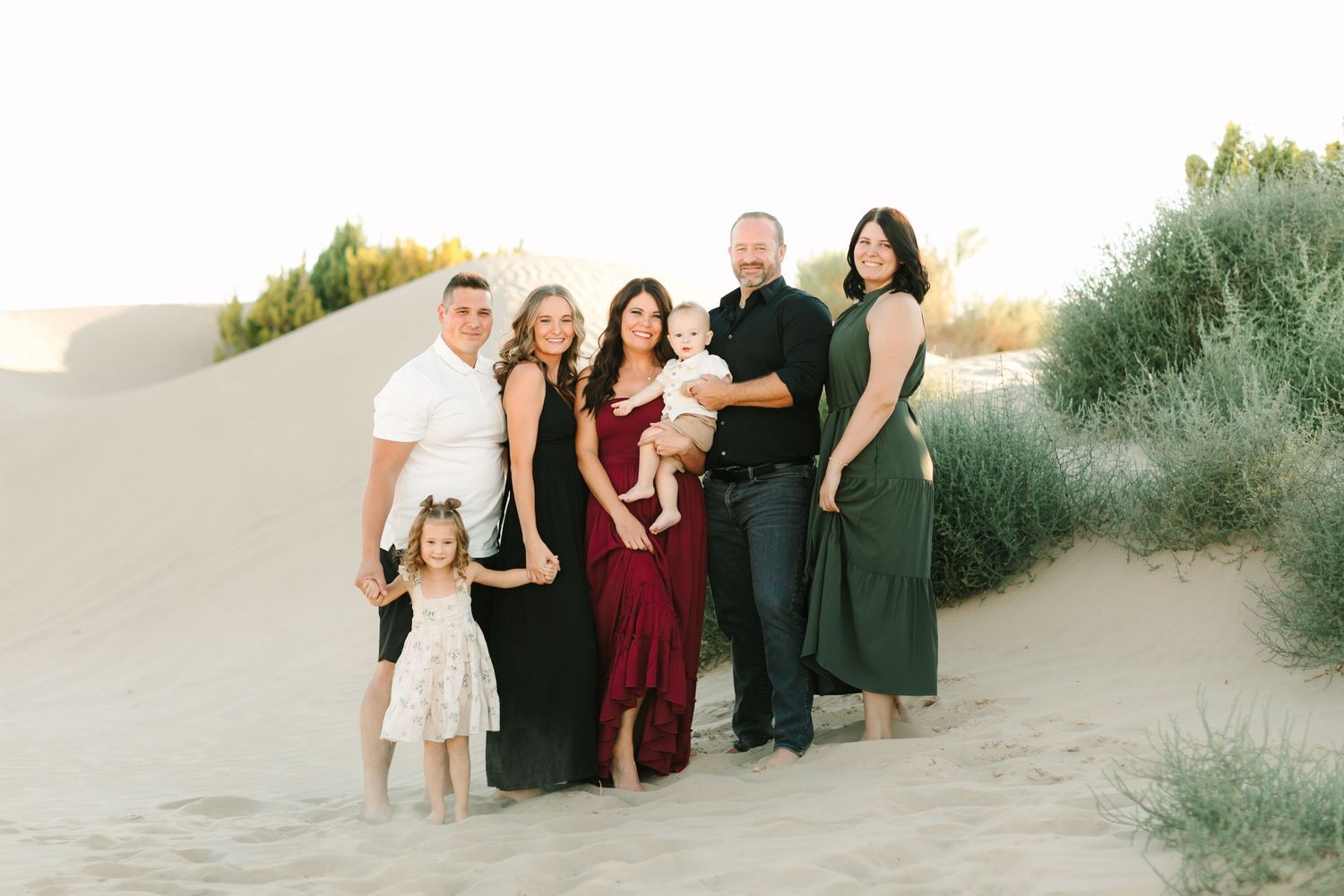A group of people in formal attire pose together on sandy dunes at sunset for outdoor photos.