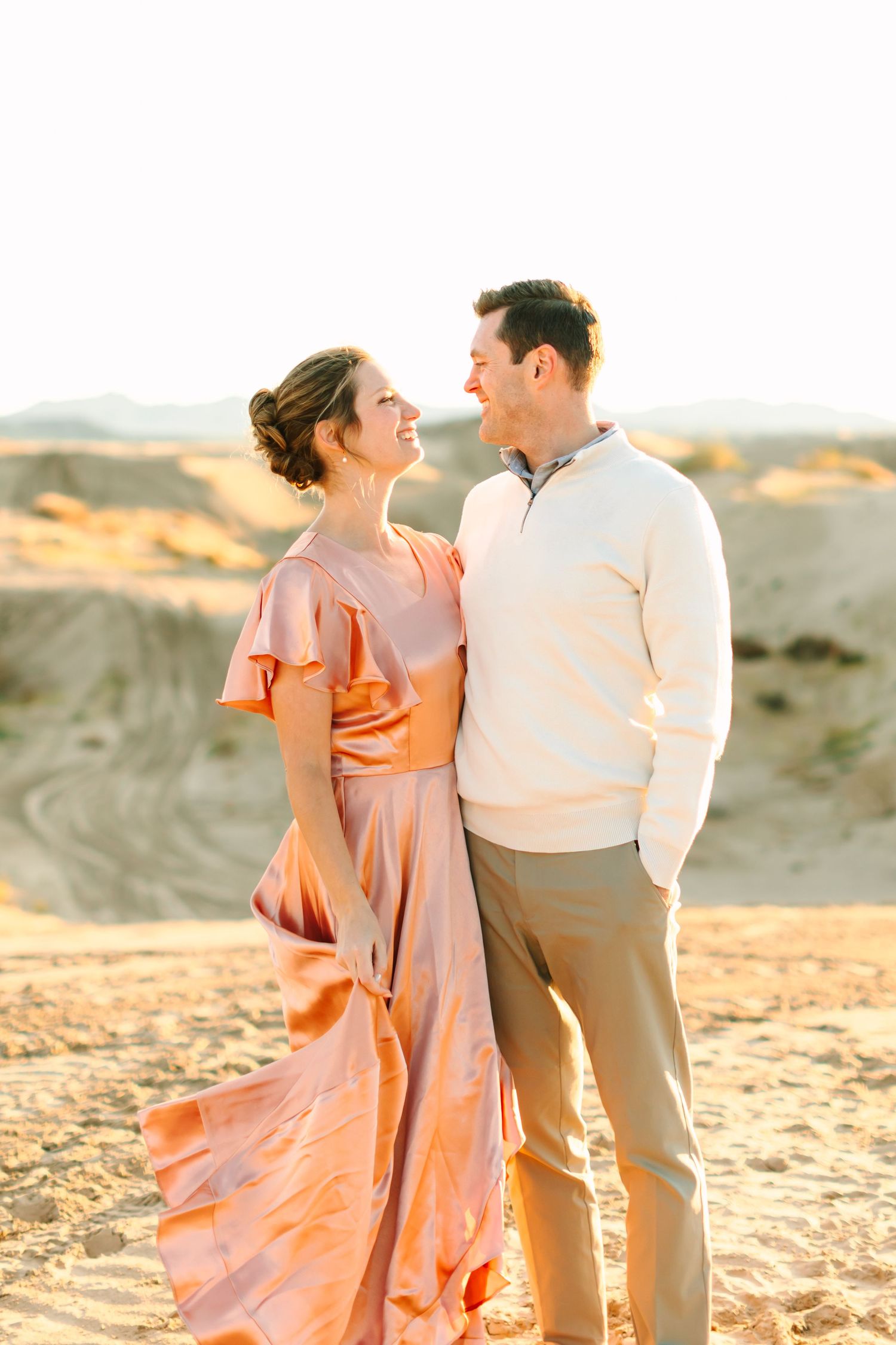 A couple in romantic desert engagement photo at sunset with woman wearing flowing peach dress and man in cream sweater.