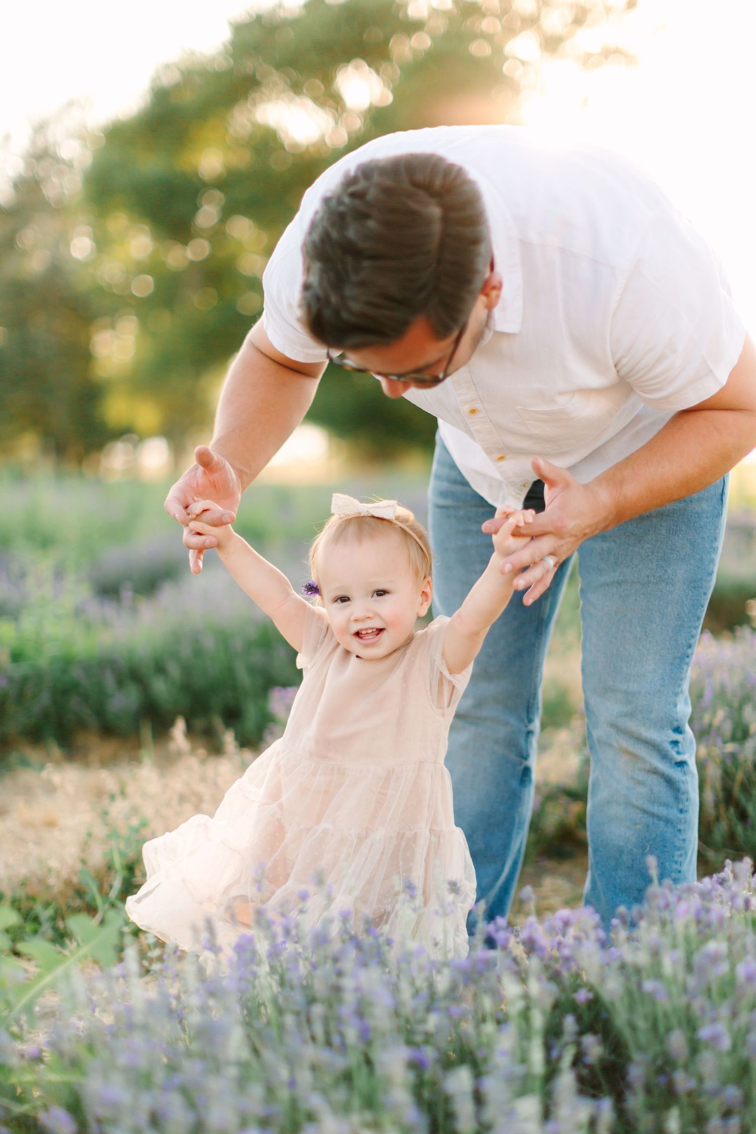 A parent in jeans and white shirt bends down to play with a small child among blooming purple lavender flowers at sunset.