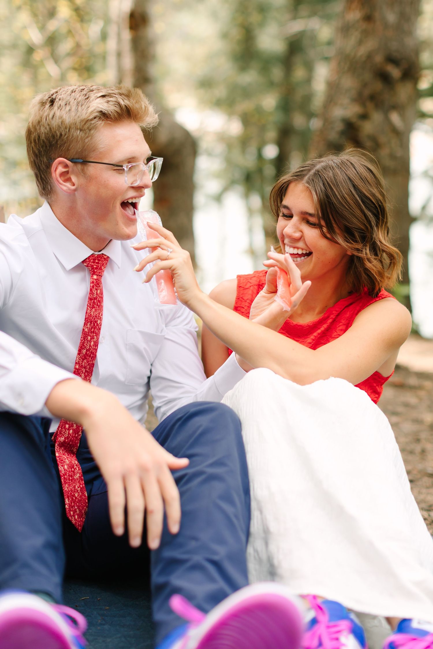 Happy couple laughing outdoors, man in white shirt and red tie, woman in red dress, sitting together in a forest setting.