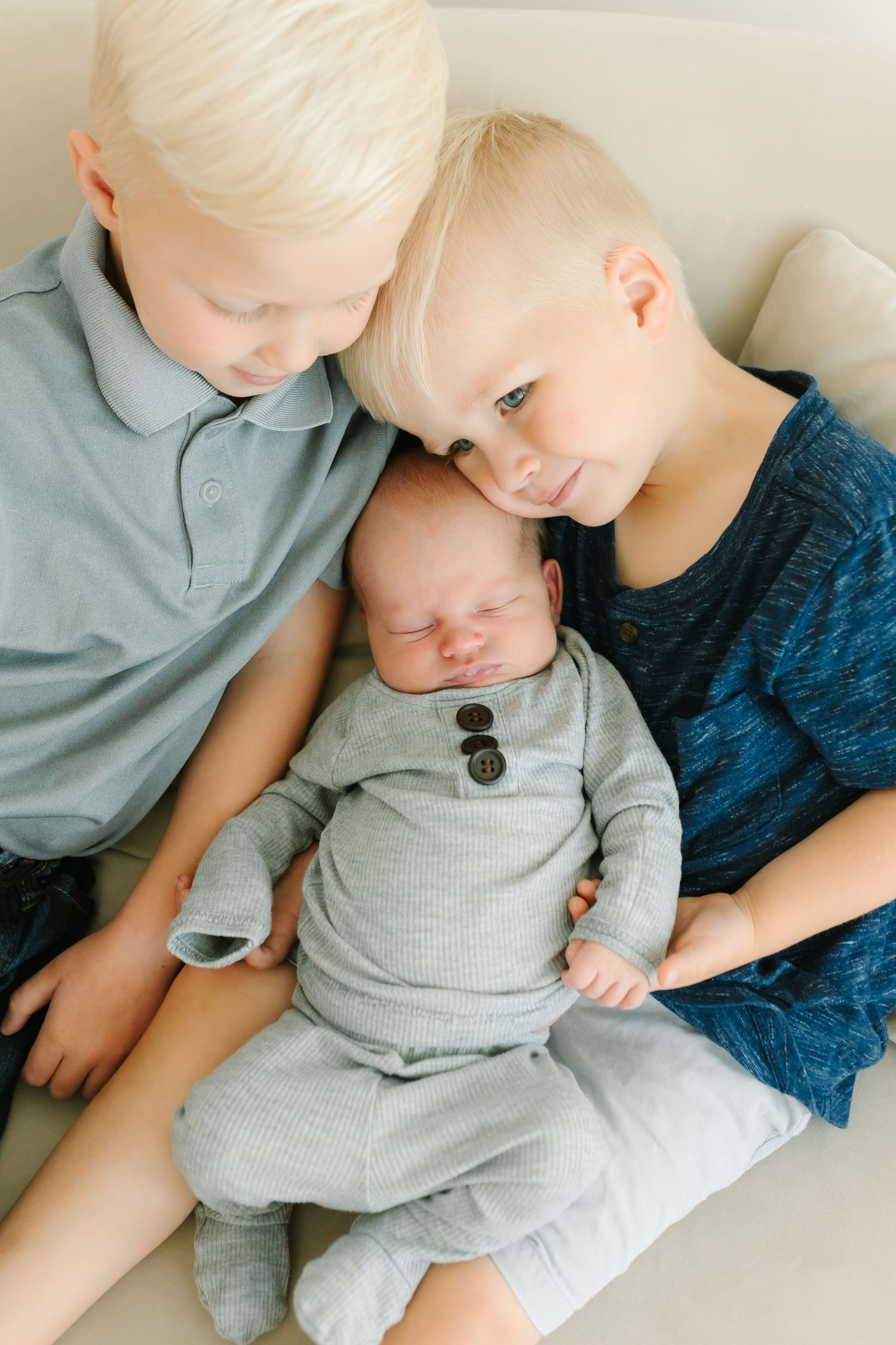 Two blonde siblings lovingly cradle their sleeping newborn baby dressed in a gray outfit on a light sofa.