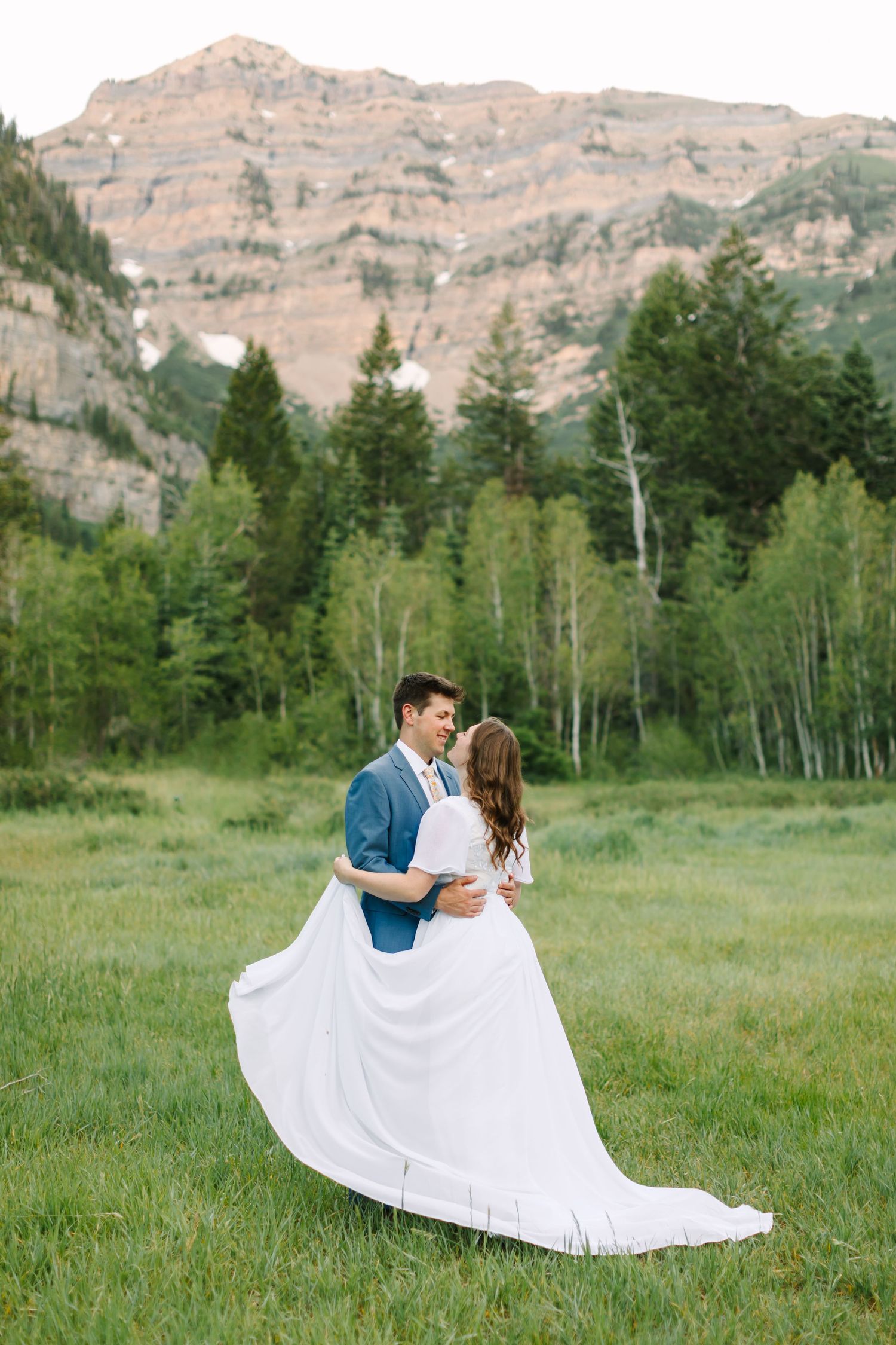 Groom lifts bride in white flowing dress while dancing in a scenic mountain meadow with rocky peaks behind them.
