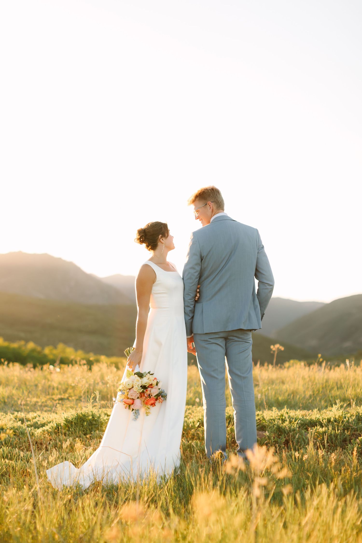 Bride and groom holding hands in a sunlit mountain meadow, bride in white gown with bouquet, groom in light blue suit.