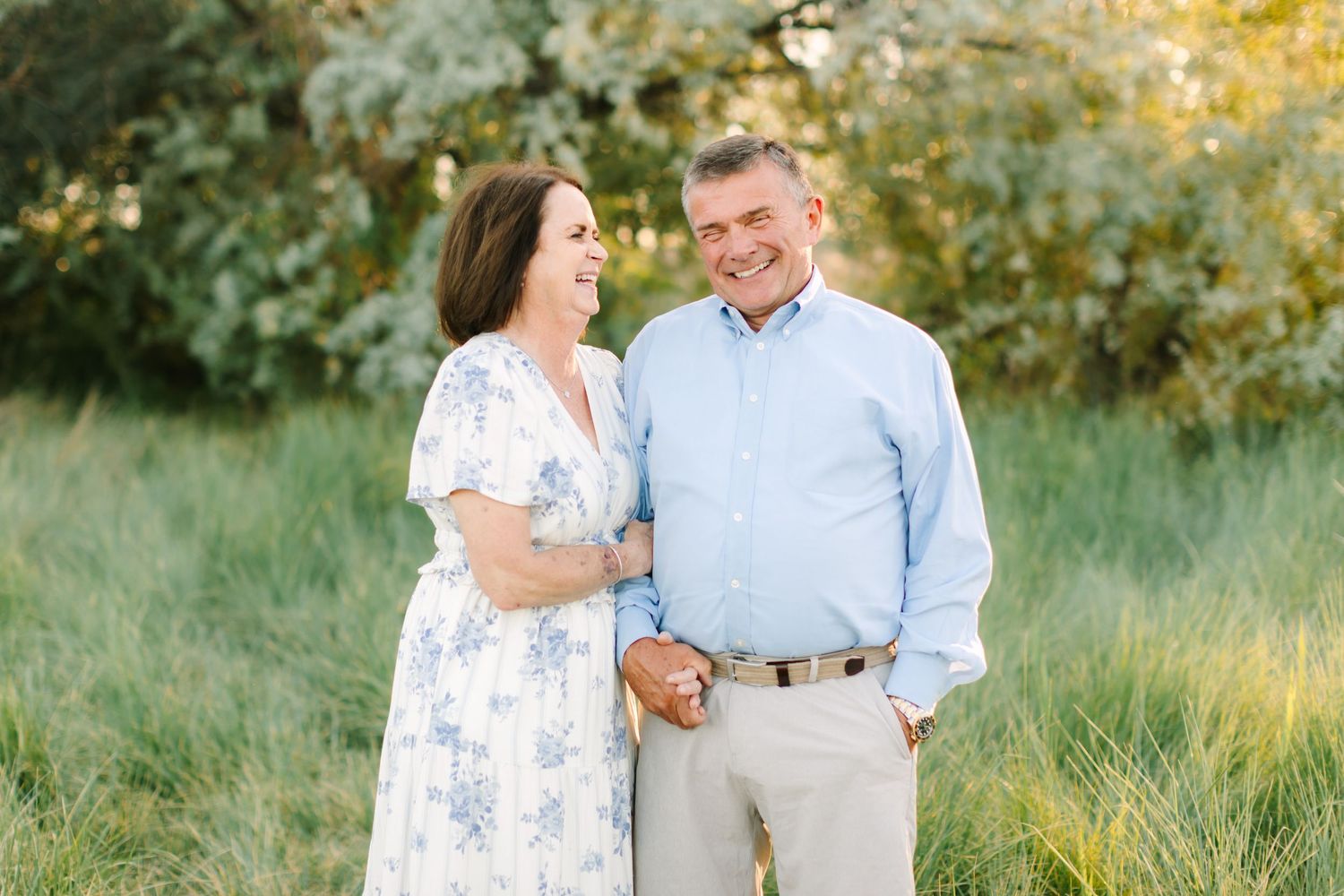 Couple laughing and holding hands in a sunlit meadow, woman in floral dress, man in blue shirt.