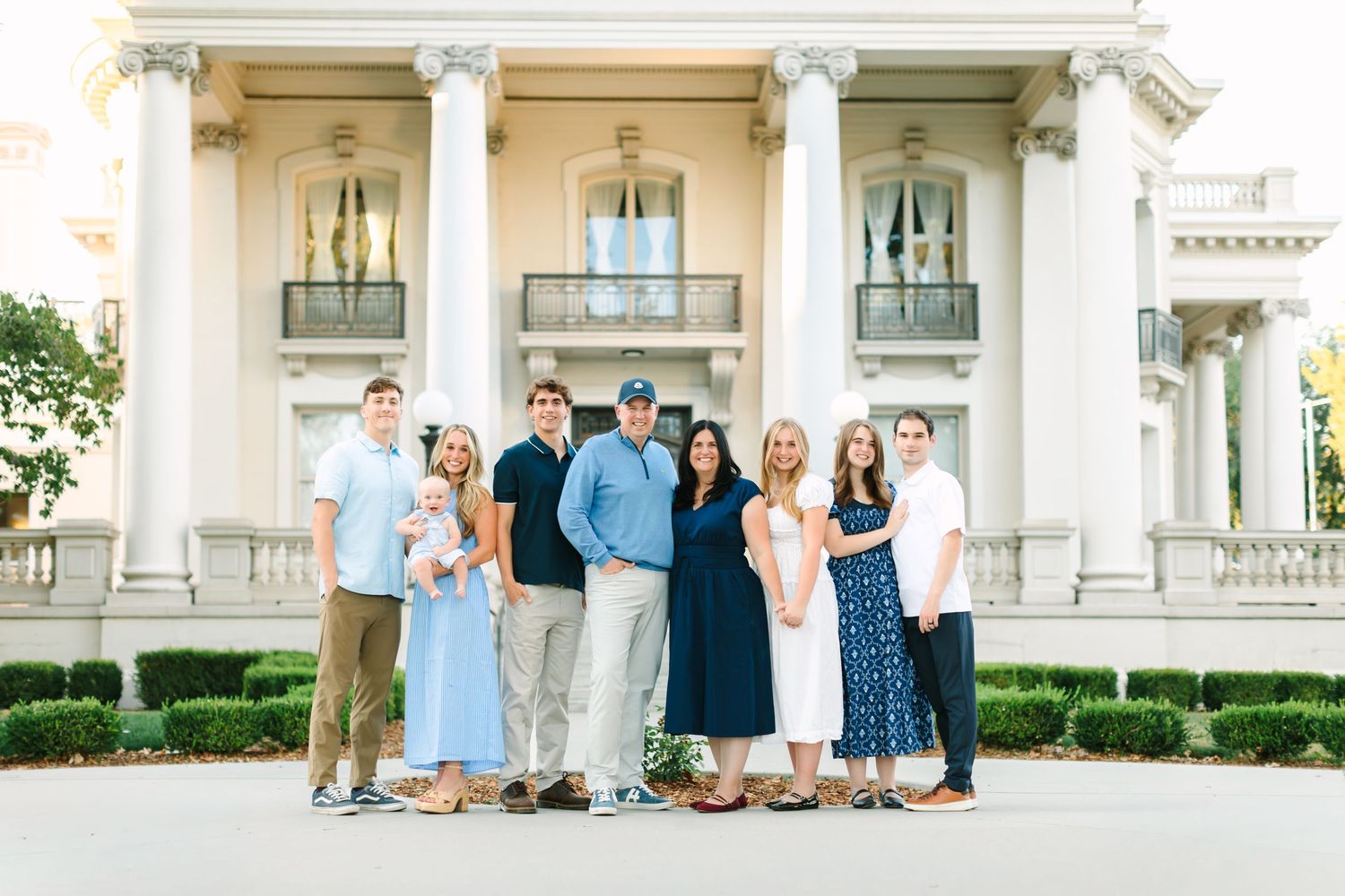 A large family poses together in front of an elegant white colonial mansion with tall column in Salt Lake City, Utah.