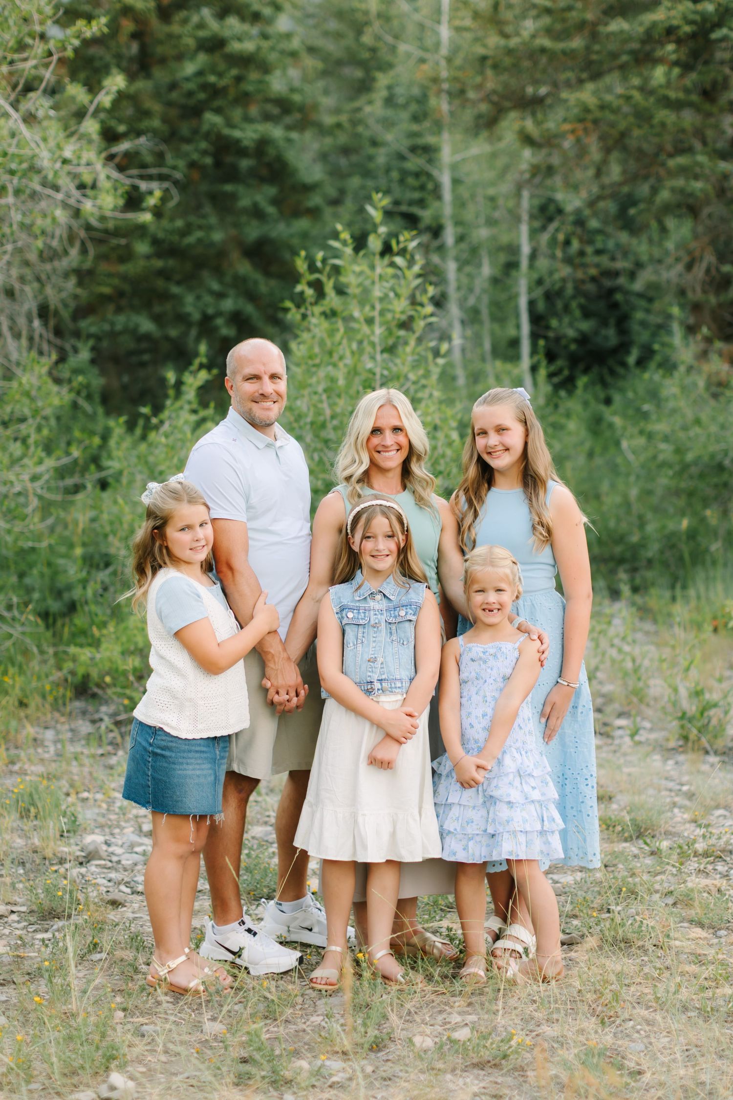 Family of six posing outdoors in coordinating pastel outfits, surrounded by lush green trees in a natural setting.