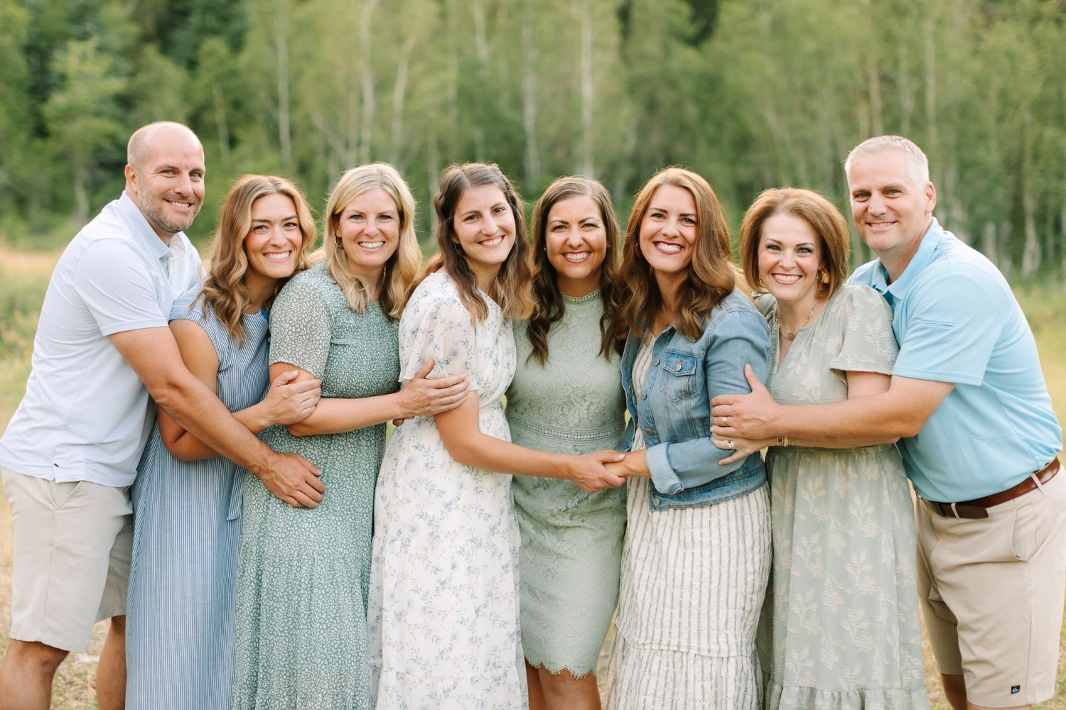 Large family group smiling outdoors in coordinated sage green and white outfits with lush green trees behind in Provo, Utah.