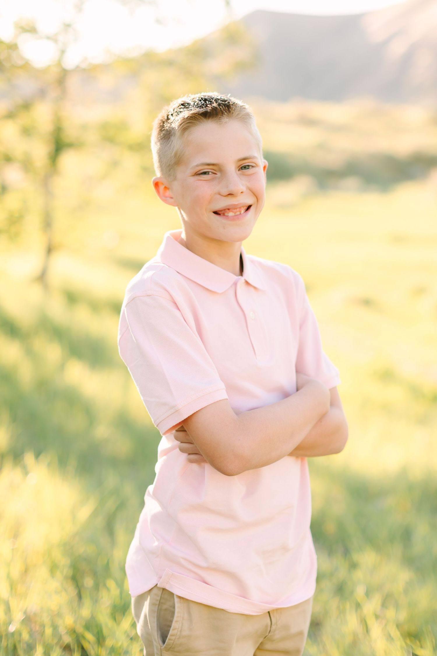 A smiling boy in a pink polo shirt stands with arms crossed in a sunny green field with mountains in the background.