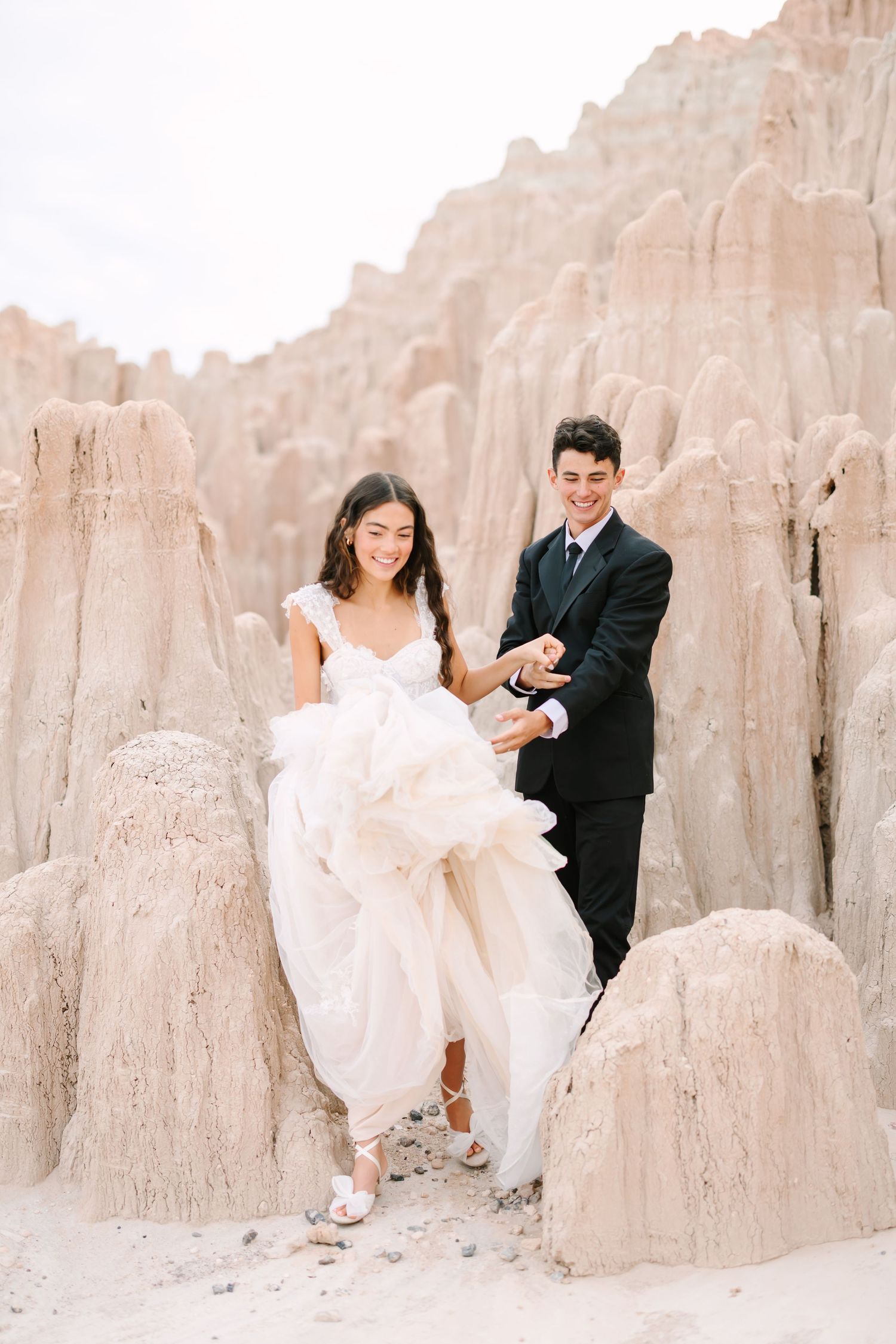 A couple in wedding attire poses among unique sand formations in a desert landscape.