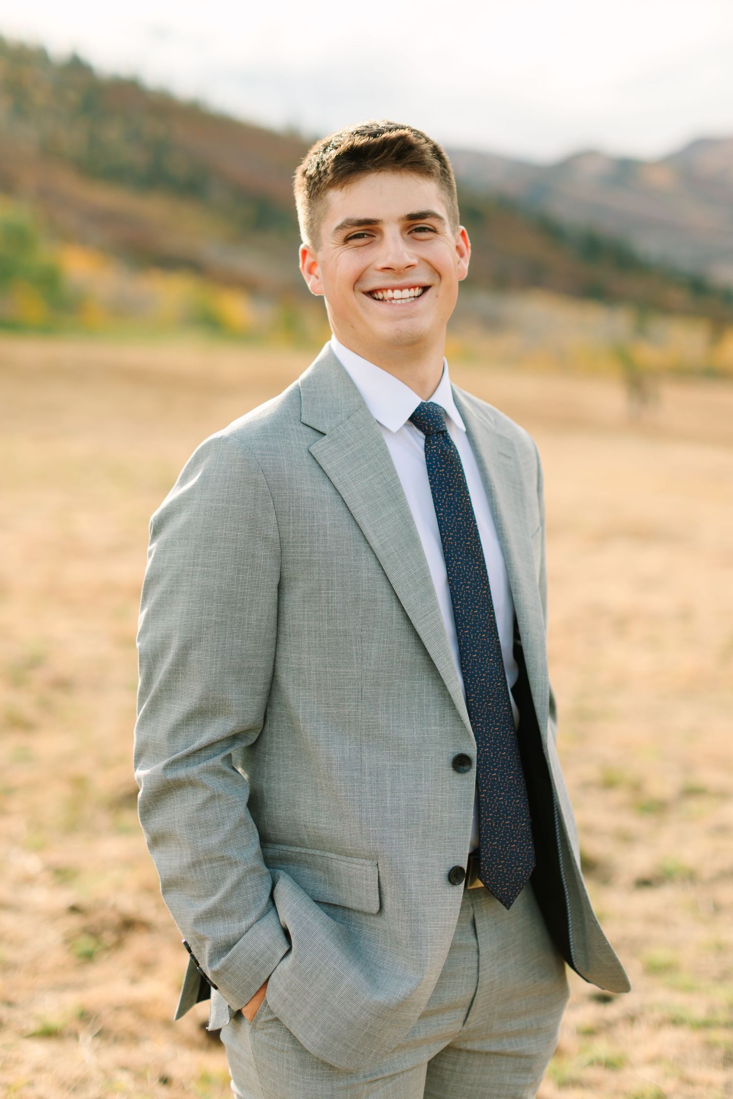Candid outdoor portrait of a person in a light gray suit smiling warmly against a mountainous backdrop.
