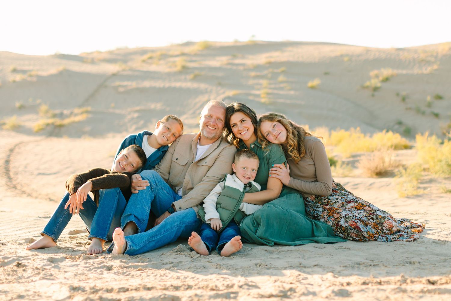 Family in coordinating blue and teal outfits sitting together on sandy hill during golden hour in desert landscape.