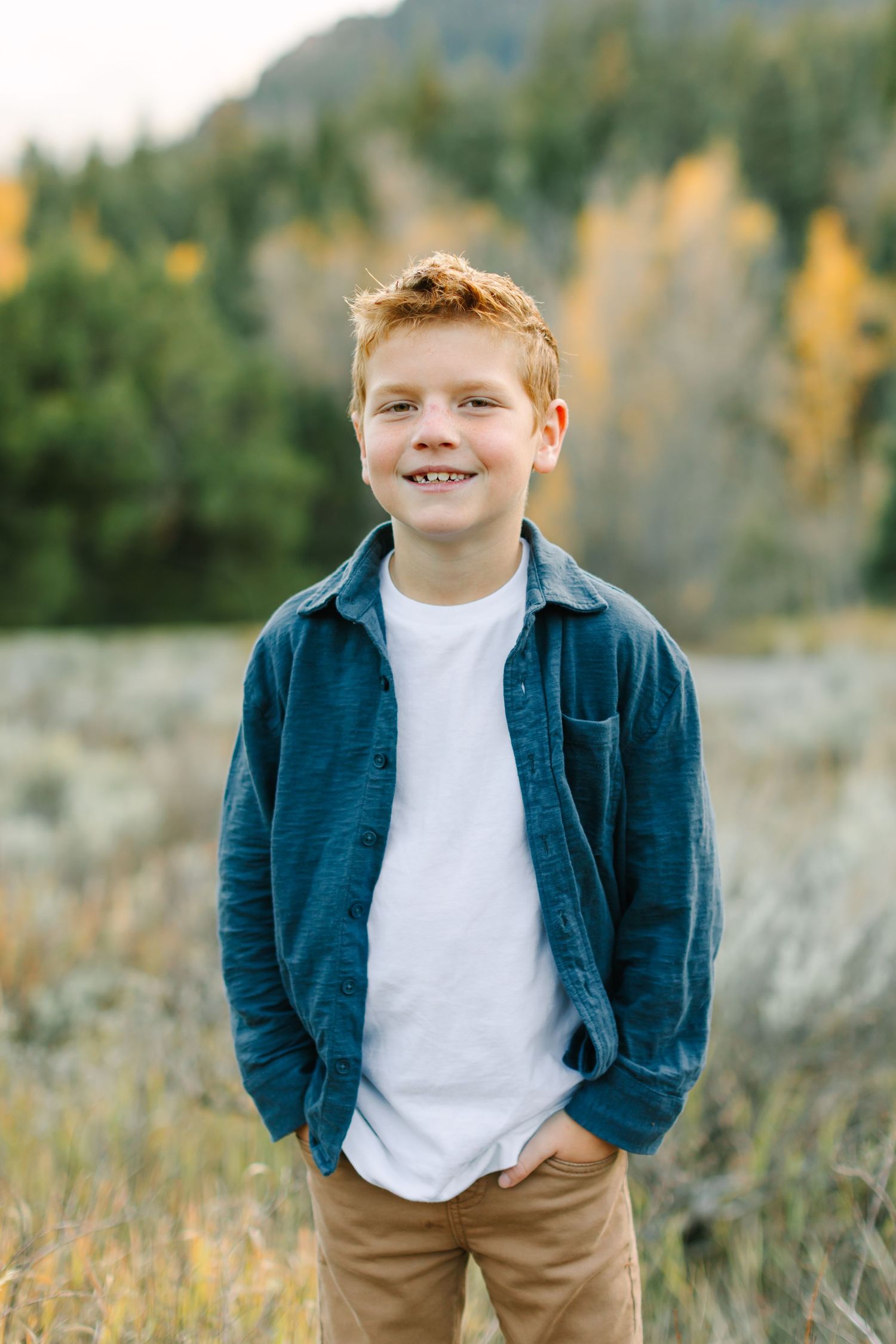A child in a navy blue jacket and khaki pants poses outdoors in a field with autumn foliage in the background.