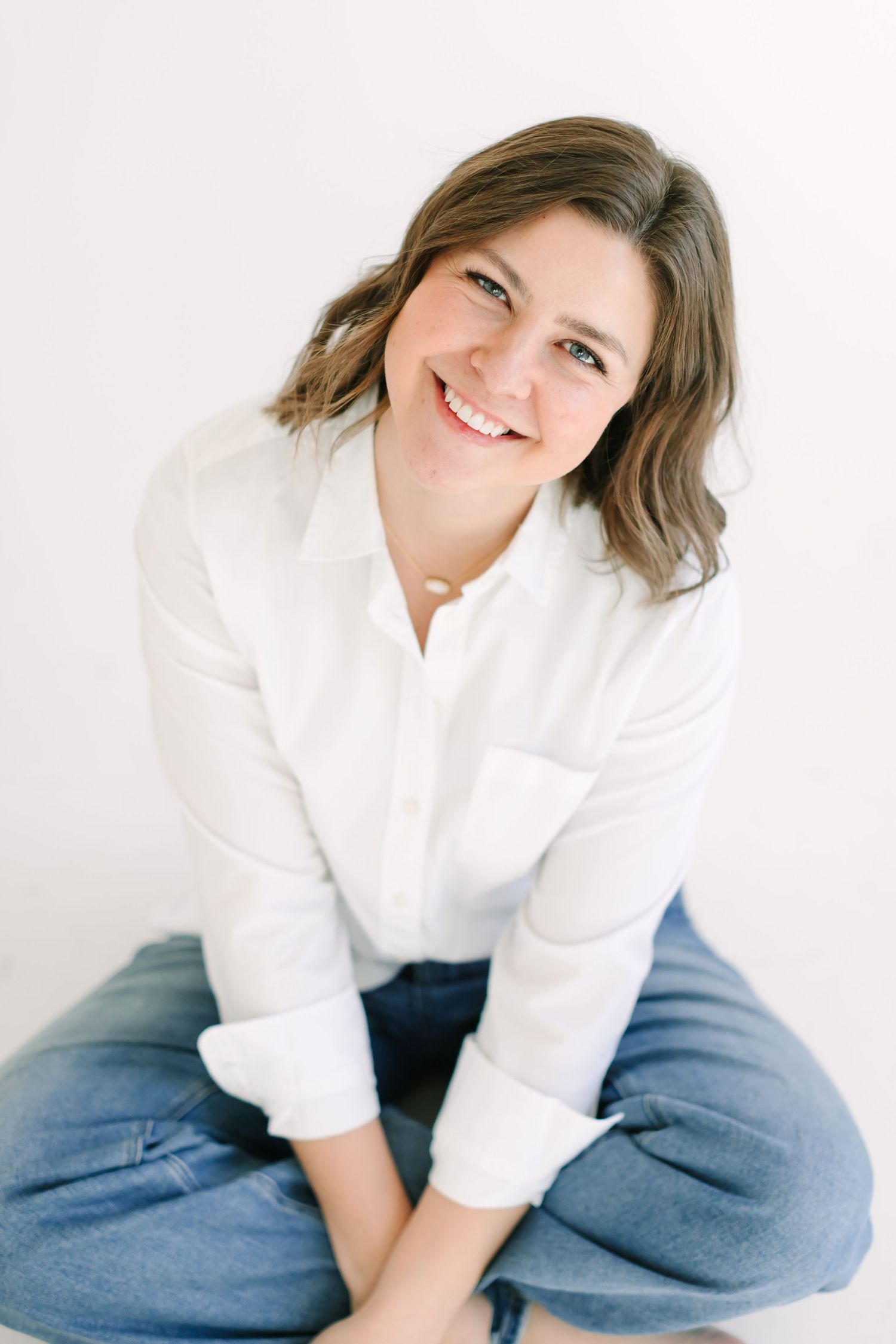 A person in a crisp white button-down shirt and blue jeans sits casually against a light background.