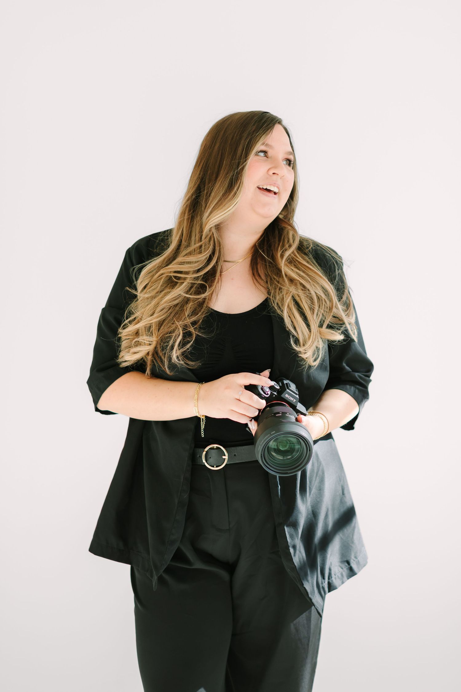 Professional photographer in black outfit holds camera while posing against white studio backdrop.