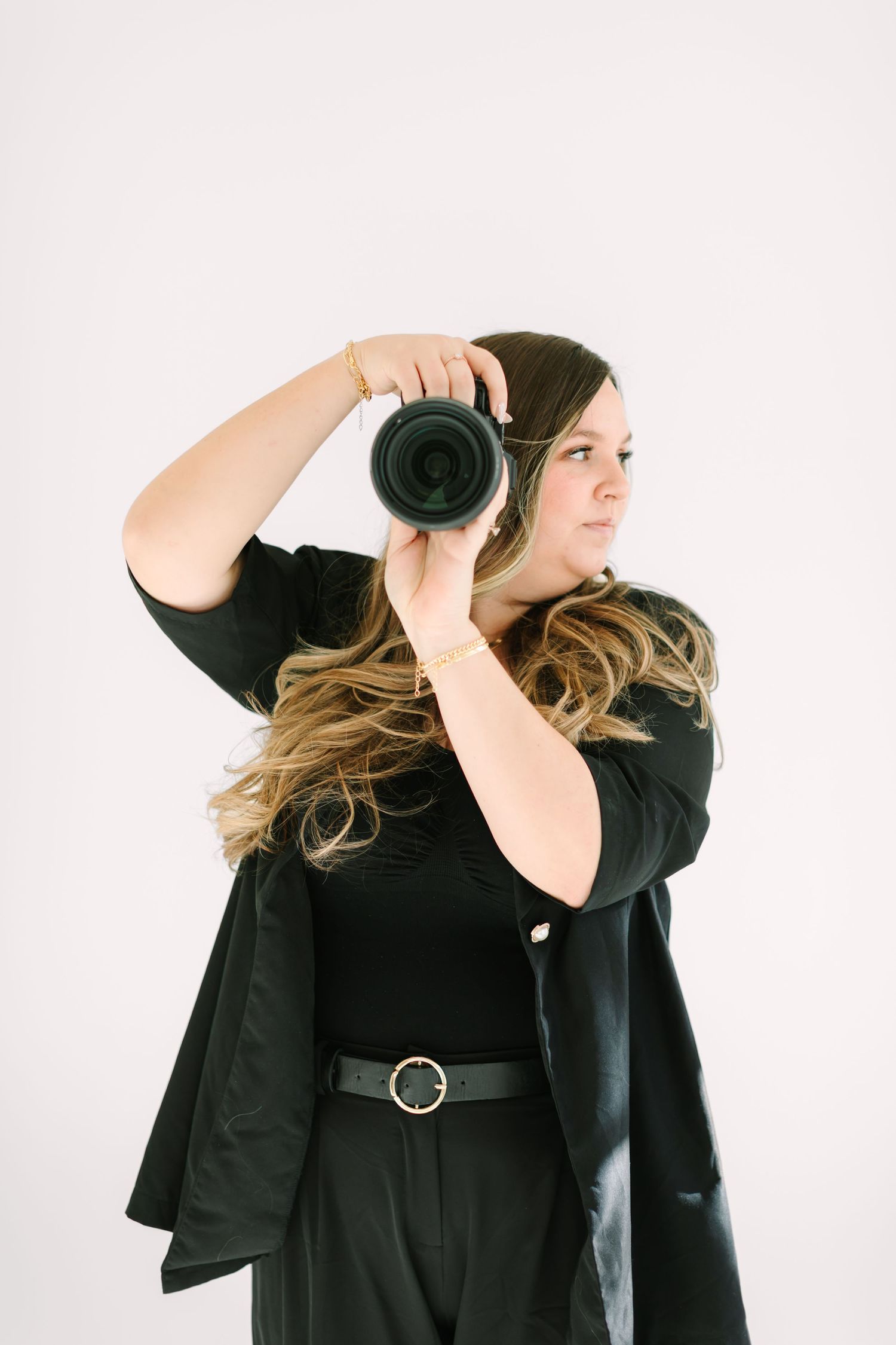 Professional photographer in black outfit with belted waist holds camera while shooting against white background.