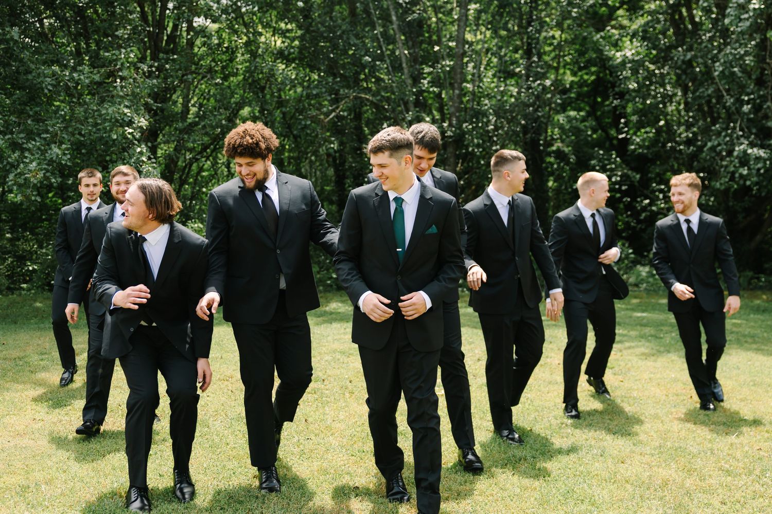 Groomsmen in black suits walking together through a lush green garden setting during a wedding celebration.