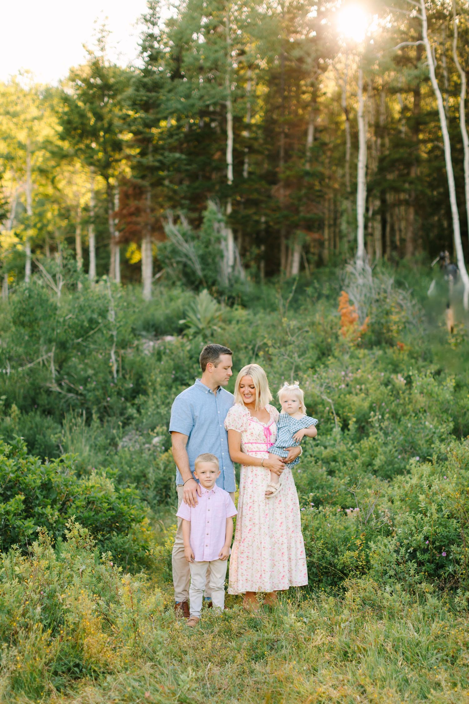 Family of four posing in a sunlit forest meadow, parents with two young children in pastel outfits surrounded by lush greenery.
