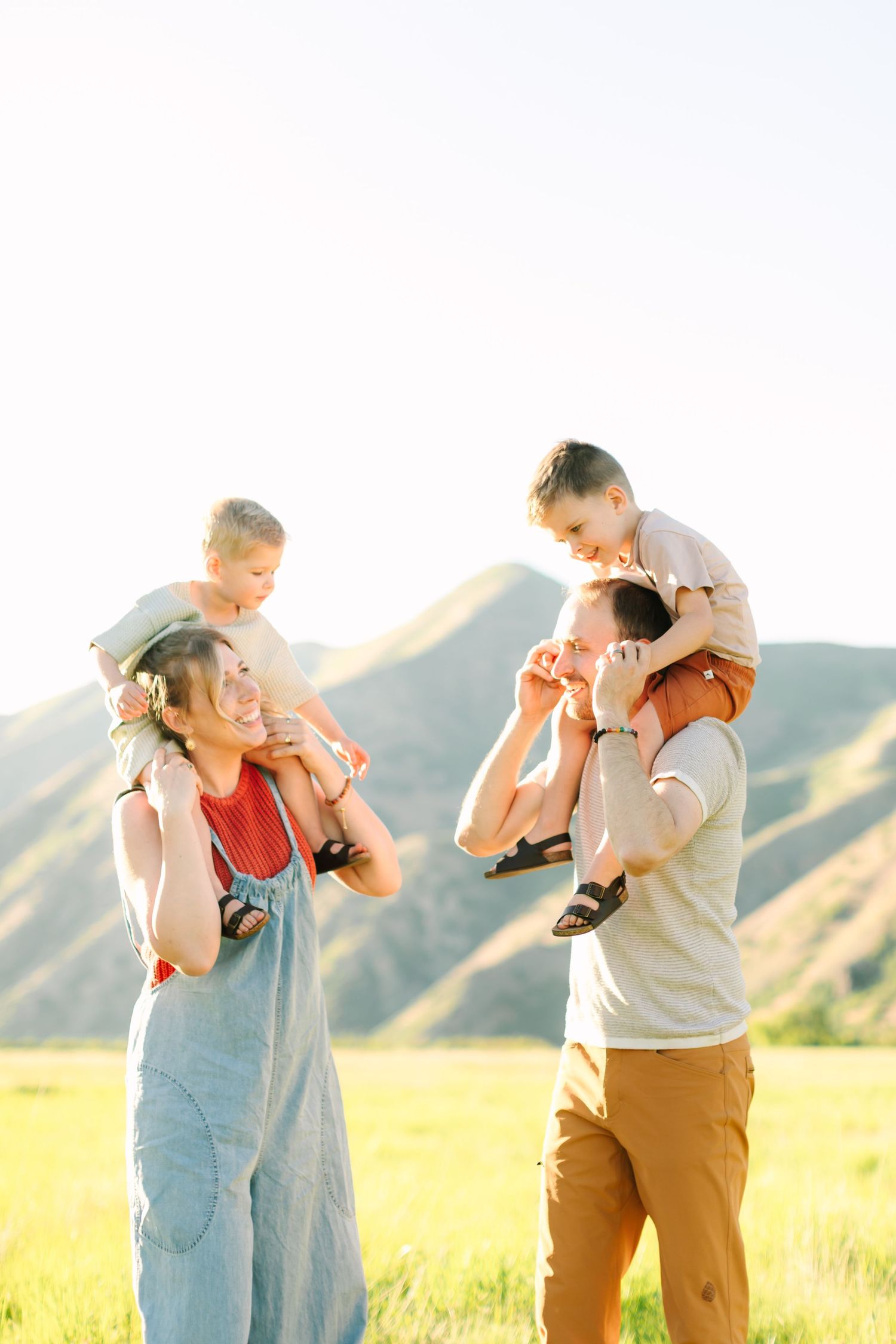 Happy family with two children on parents shoulders in a sunny green meadow with mountain backdrop.