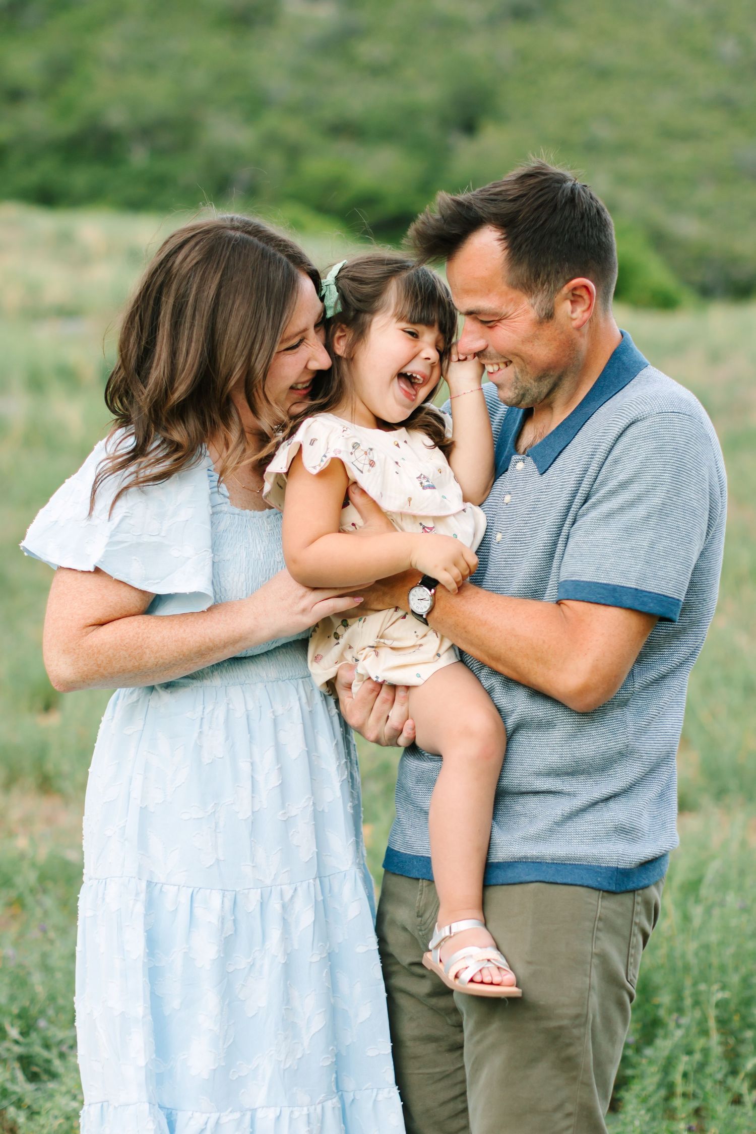 A joyful family of three poses outdoors, with parents holding their laughing toddler girl in a green meadow.