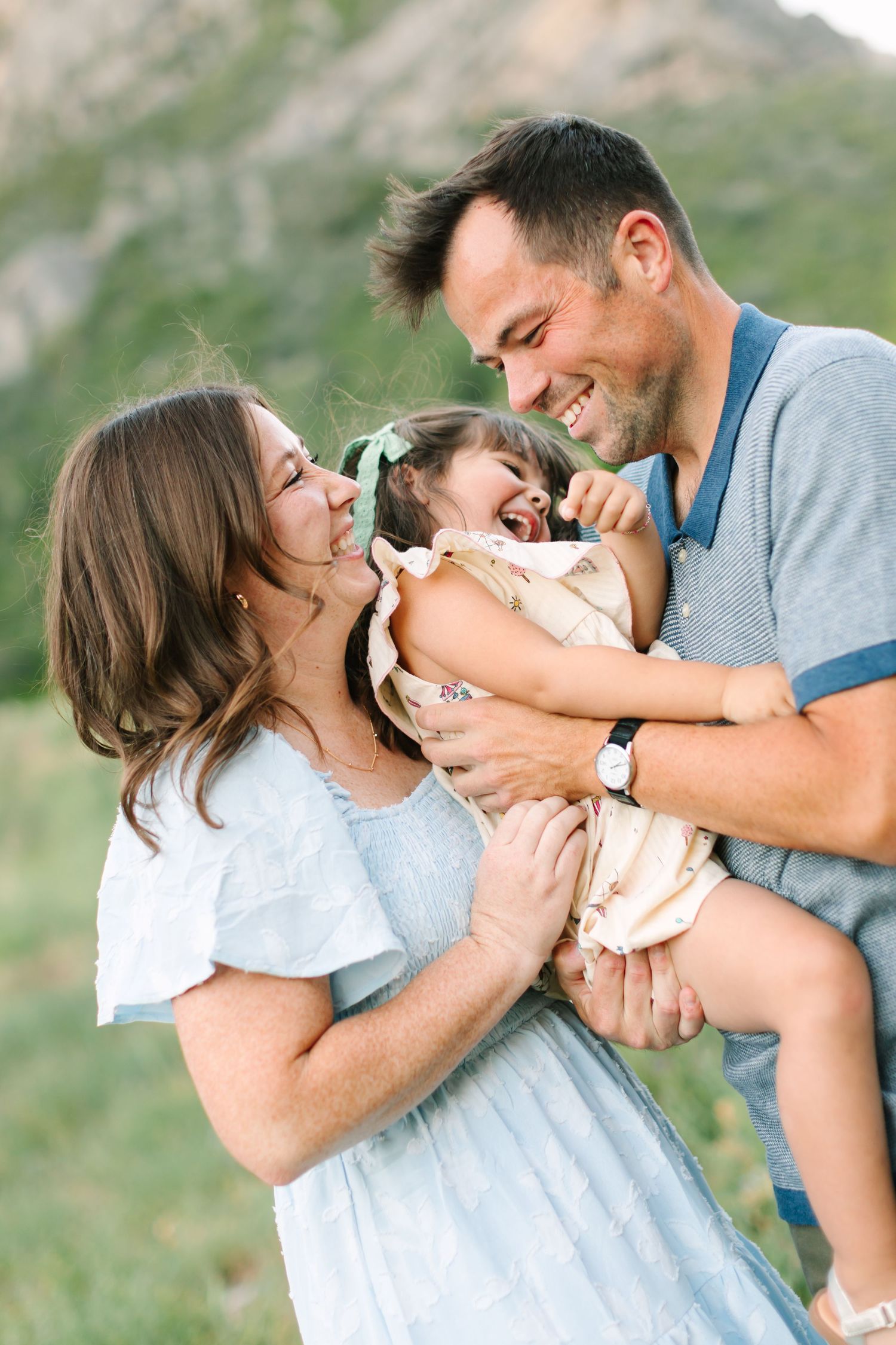 Happy family with laughing baby girl outdoors in mountain meadow, parents smiling and holding child in warm natural light.