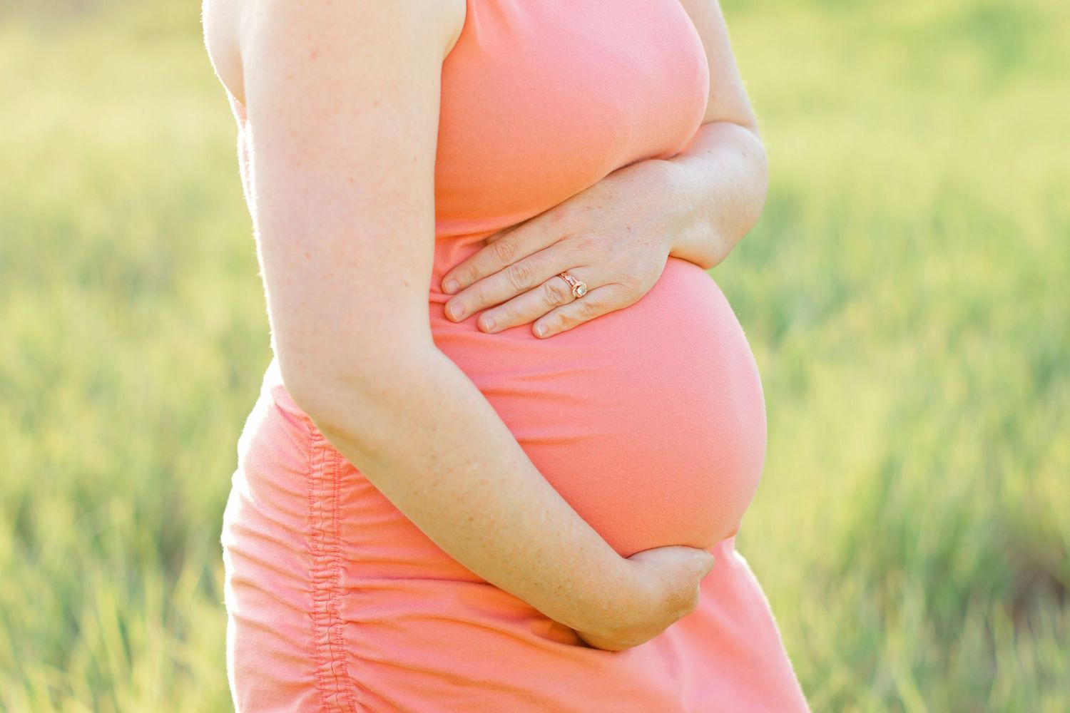 A pregnant woman in a coral dress cradles her baby bump while standing in a sunlit grassy field.