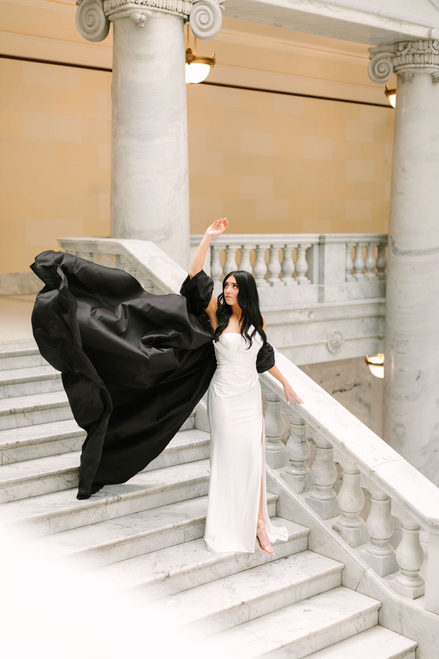 Woman in white dress poses dramatically on marble stairs with flowing black fabric creating artistic movement.