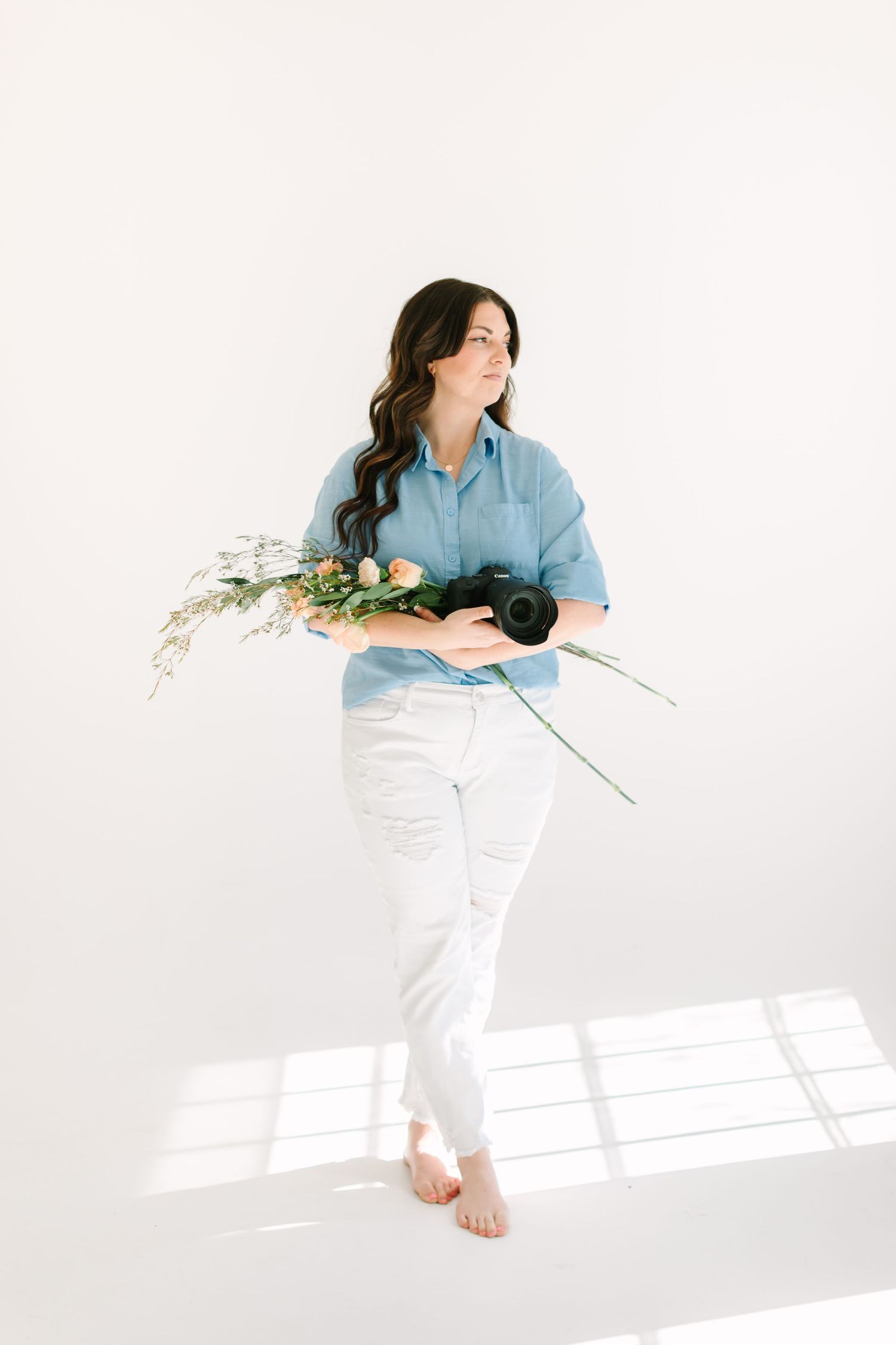 Person in blue shirt and white pants lying on floor with camera and flowers in minimalist white setting.
